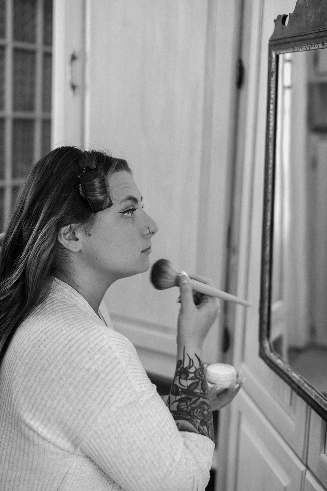 Black and white image of a bride doing her makeup in front of a mirror with rollers in her hair.