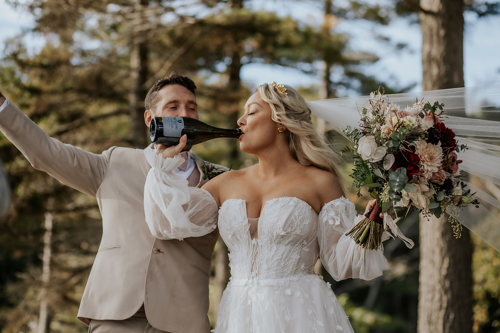 Bride celebrating and drinking from a champagne bottle holding a fall floral bouquet.
