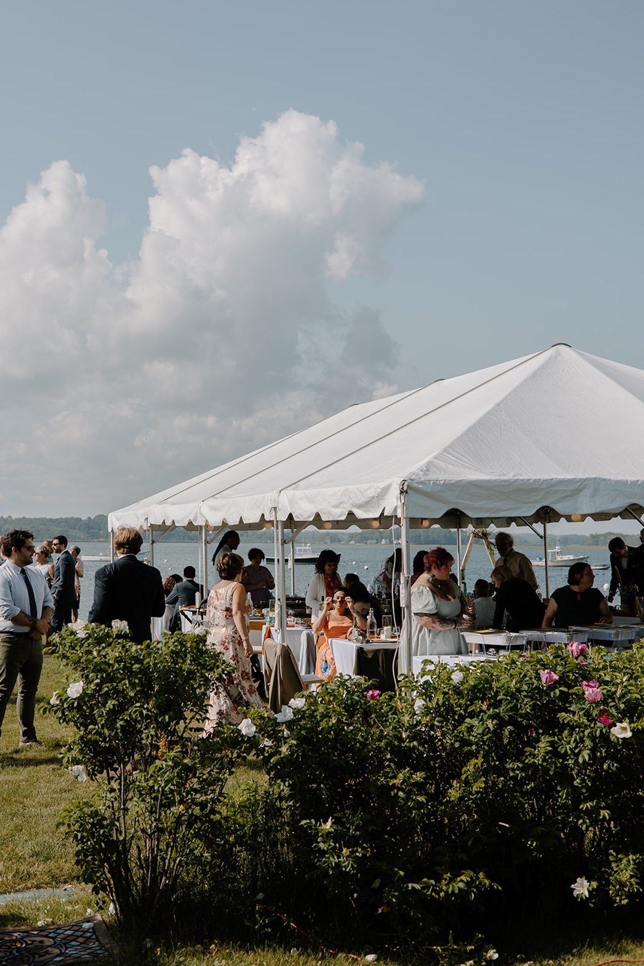 Guests mingling under a white tent by the ocean during an outdoor wedding reception with stunning waterfront views.