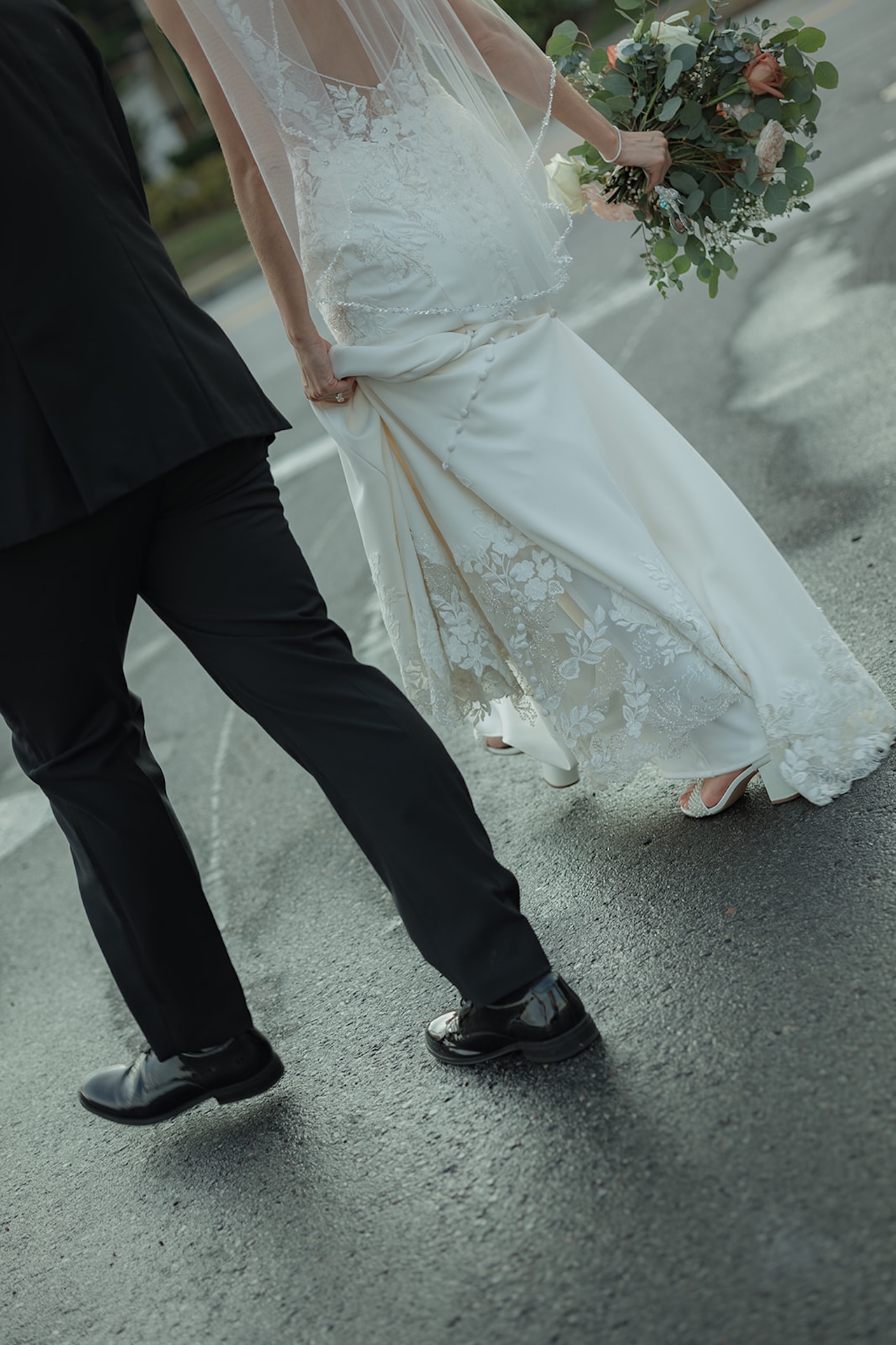 Bride and groom walking together, her lace dress trailing while she carries her bouquet, candid street-style wedding photo.