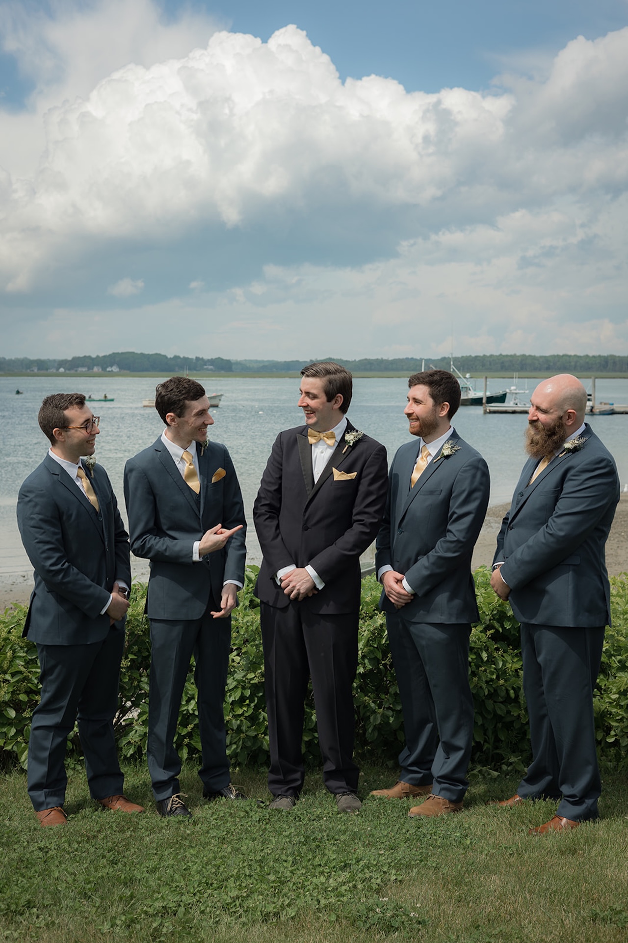Groom standing with groomsmen by the waterfront in navy suits and gold ties, laughing together before the ceremony.