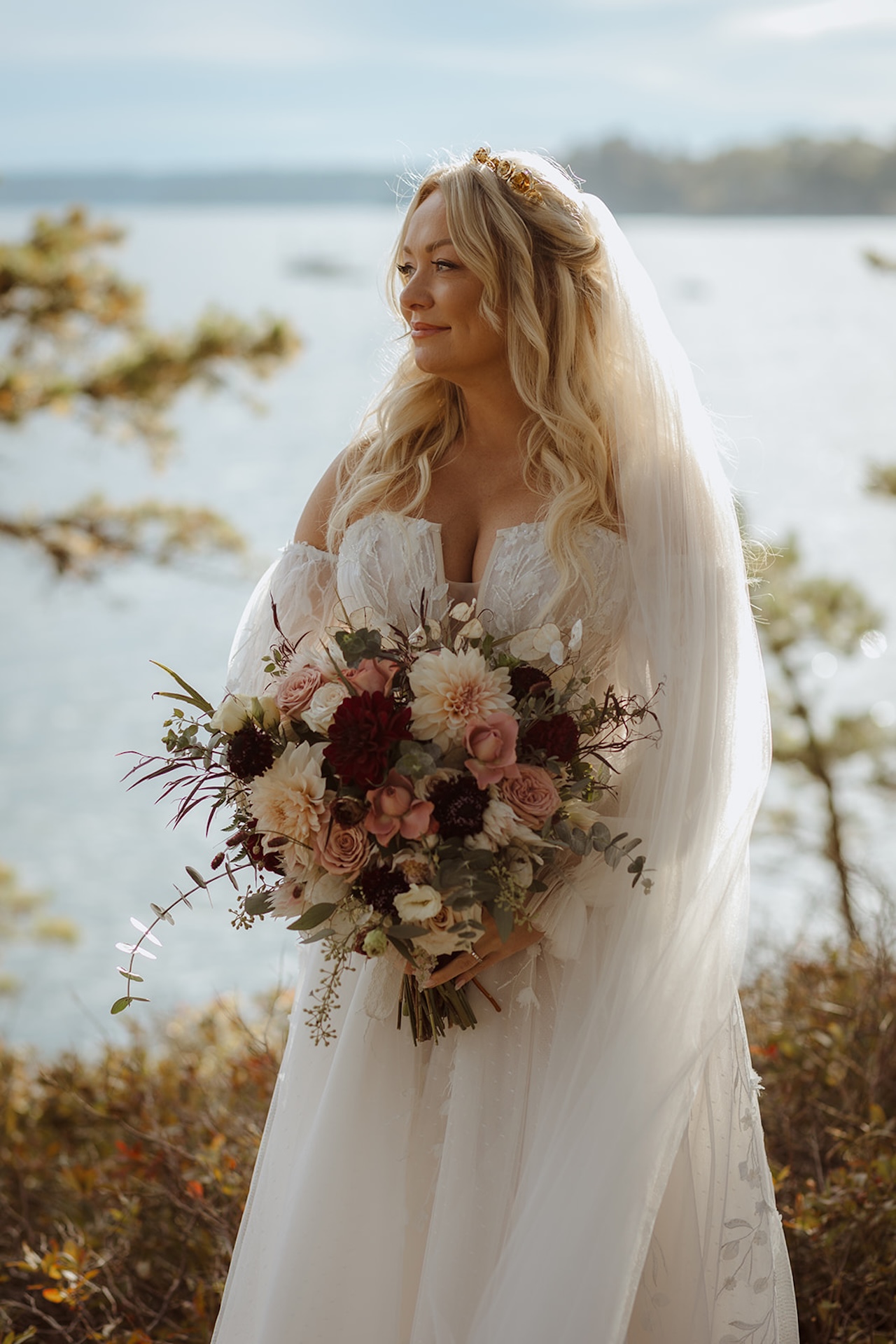 Bride with long blonde hair in an off-shoulder gown holding a large bouquet of blush and burgundy florals, a romantic capture for Destination Wedding Location Ideas.