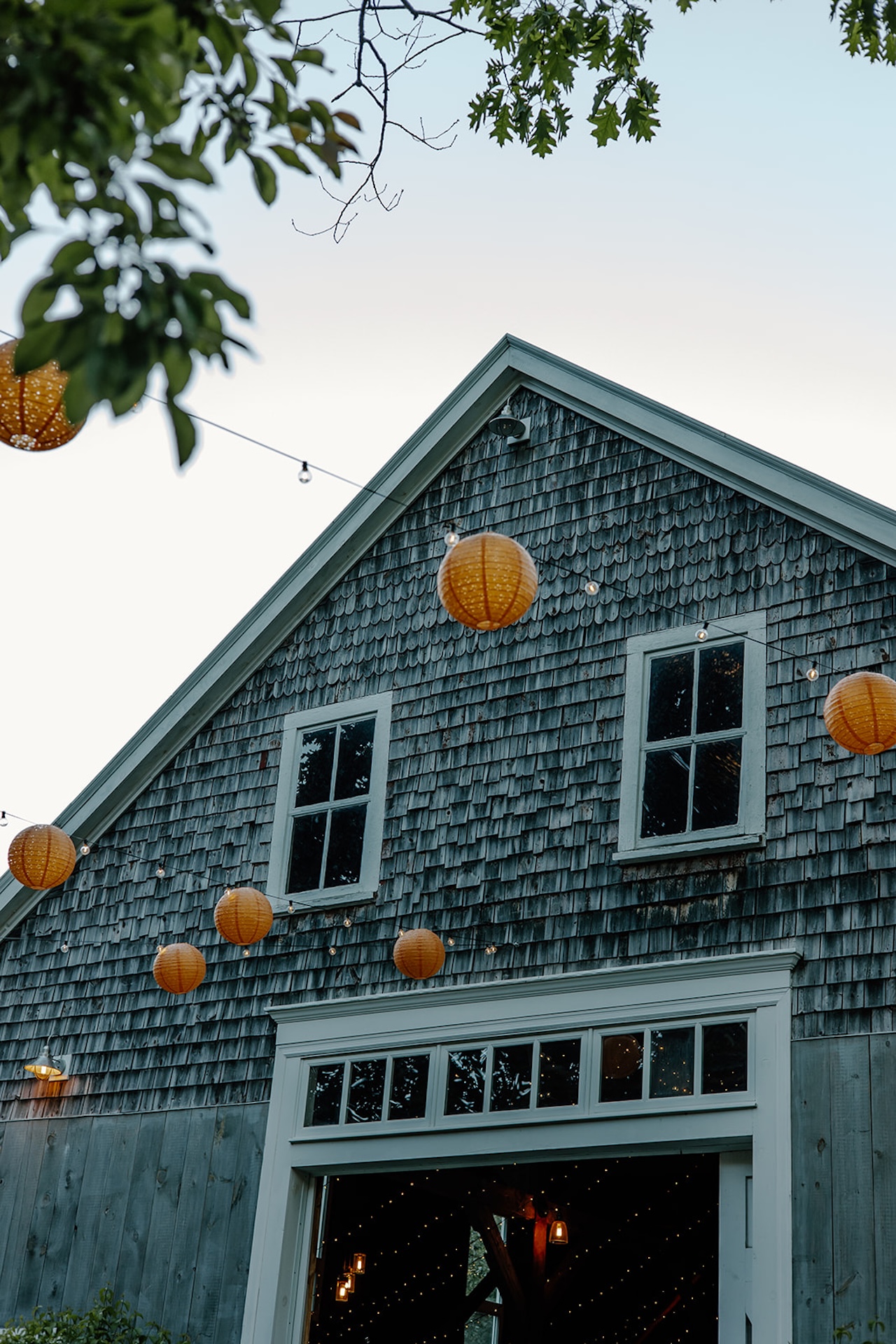 Rustic barn with weathered shingles decorated with string lights and hanging lanterns, glowing against the evening sky.