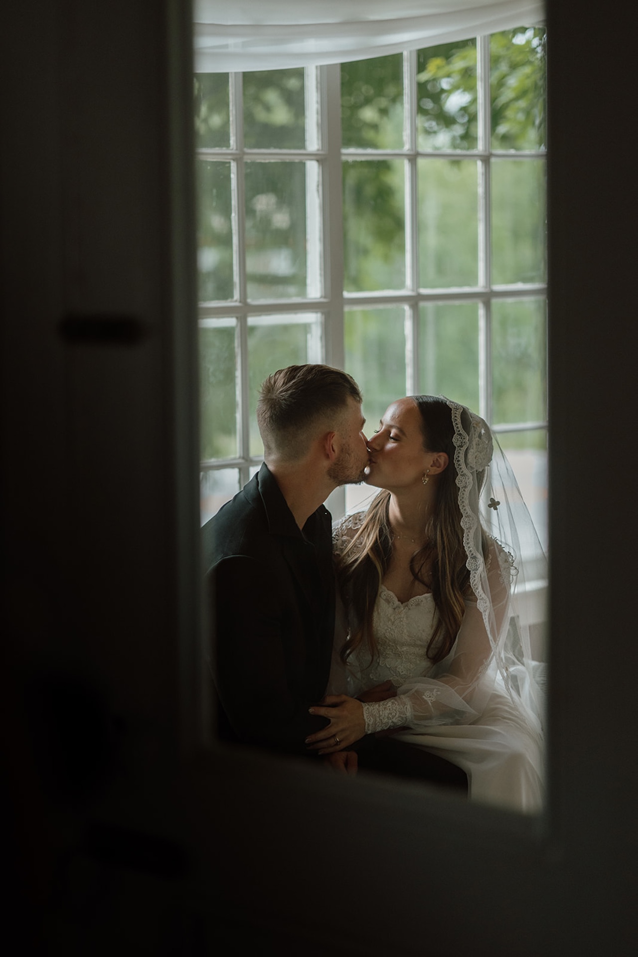 Couple sharing a kiss by a window, captured through the frame for an intimate and moody wedding portrait that highlights Destination Wedding Location Ideas.