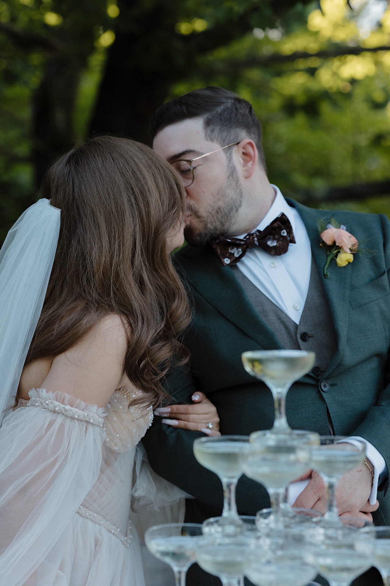 Bride and groom kissing in front of a champagne tower outdoors, her whimsical off-shoulder gown paired with his dark green suit.