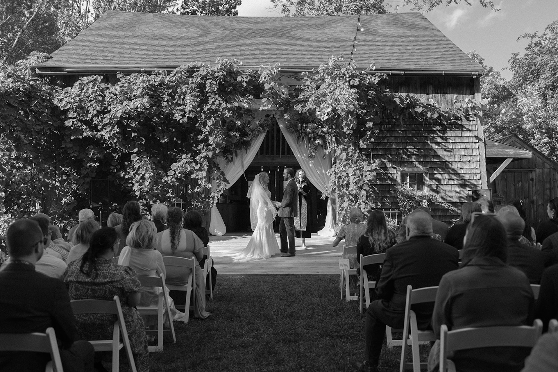 Outdoor barn wedding ceremony with draped greenery, bride and groom exchanging vows in front of gathered guests.