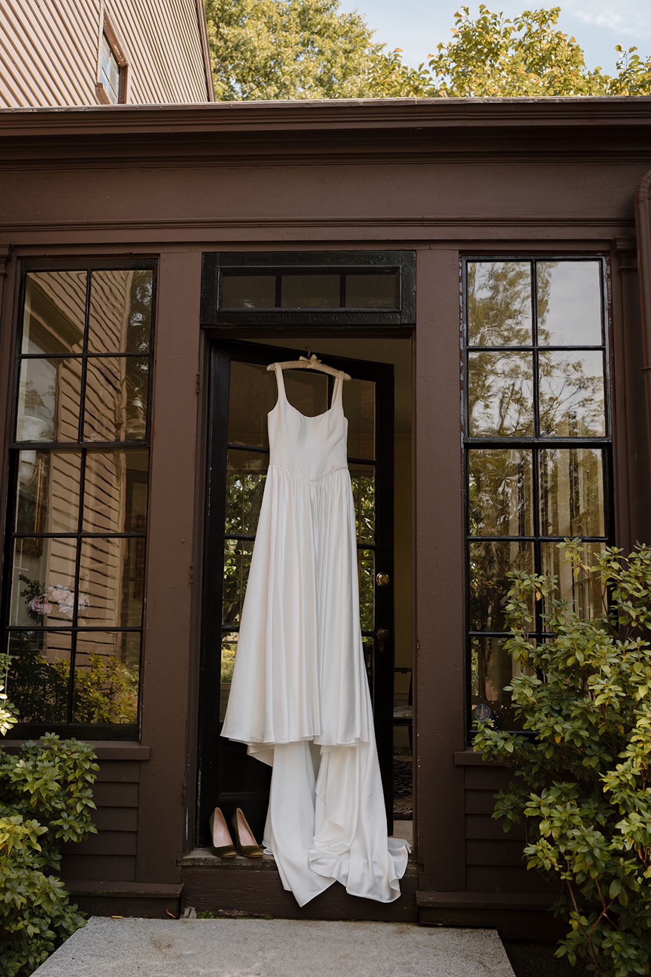 Simple white wedding dress hanging in the doorway of a rustic brown home with large glass windows, shoes placed neatly below.