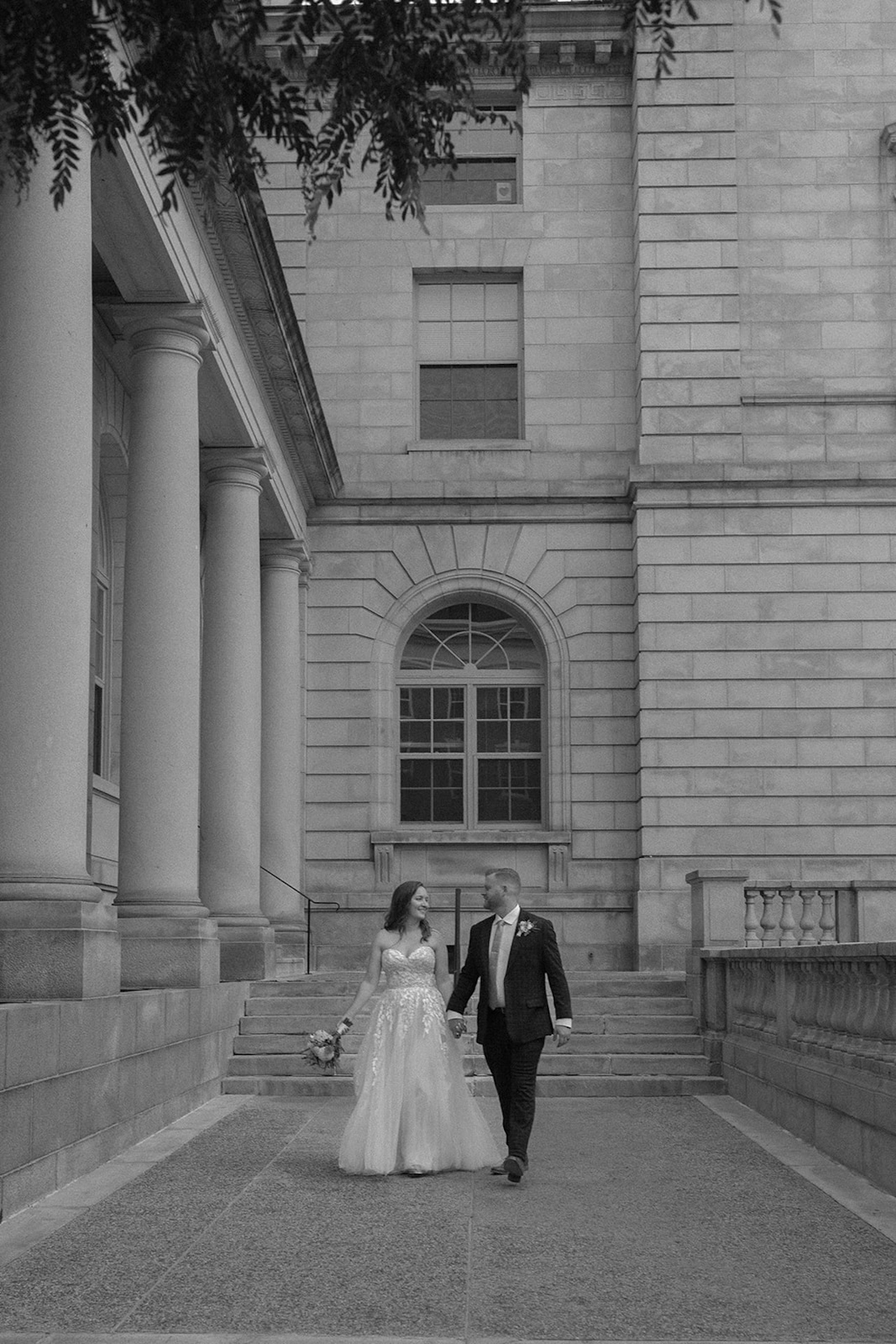 Black-and-white photo of a couple walking hand in hand through a historic stone building with columns, timeless architecture.