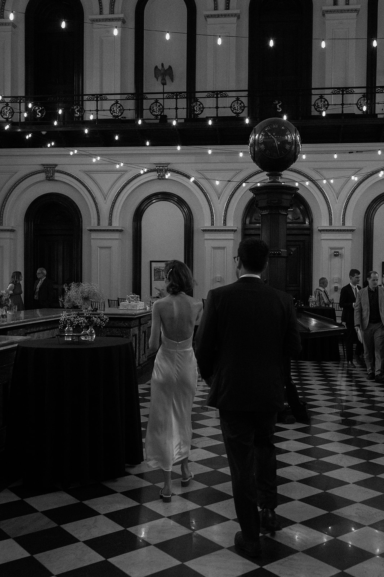 Black-and-white photo of a couple entering a grand reception hall with checkerboard floors, string lights overhead, and arched details tied to Destination Wedding Location Ideas.