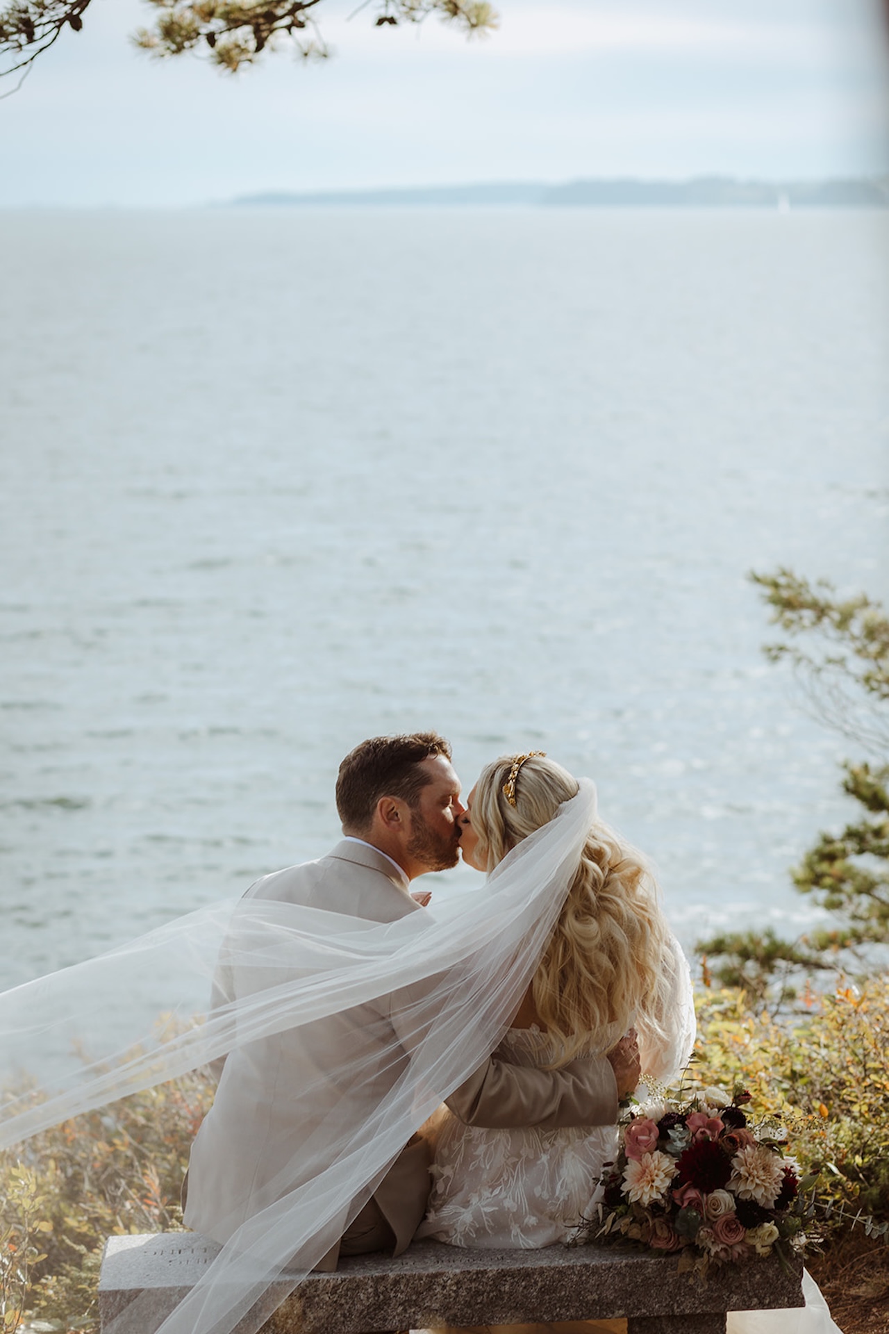 Bride and groom kissing on a stone bench overlooking the ocean, her veil flowing in the breeze, a romantic capture of Destination Wedding Location Ideas.