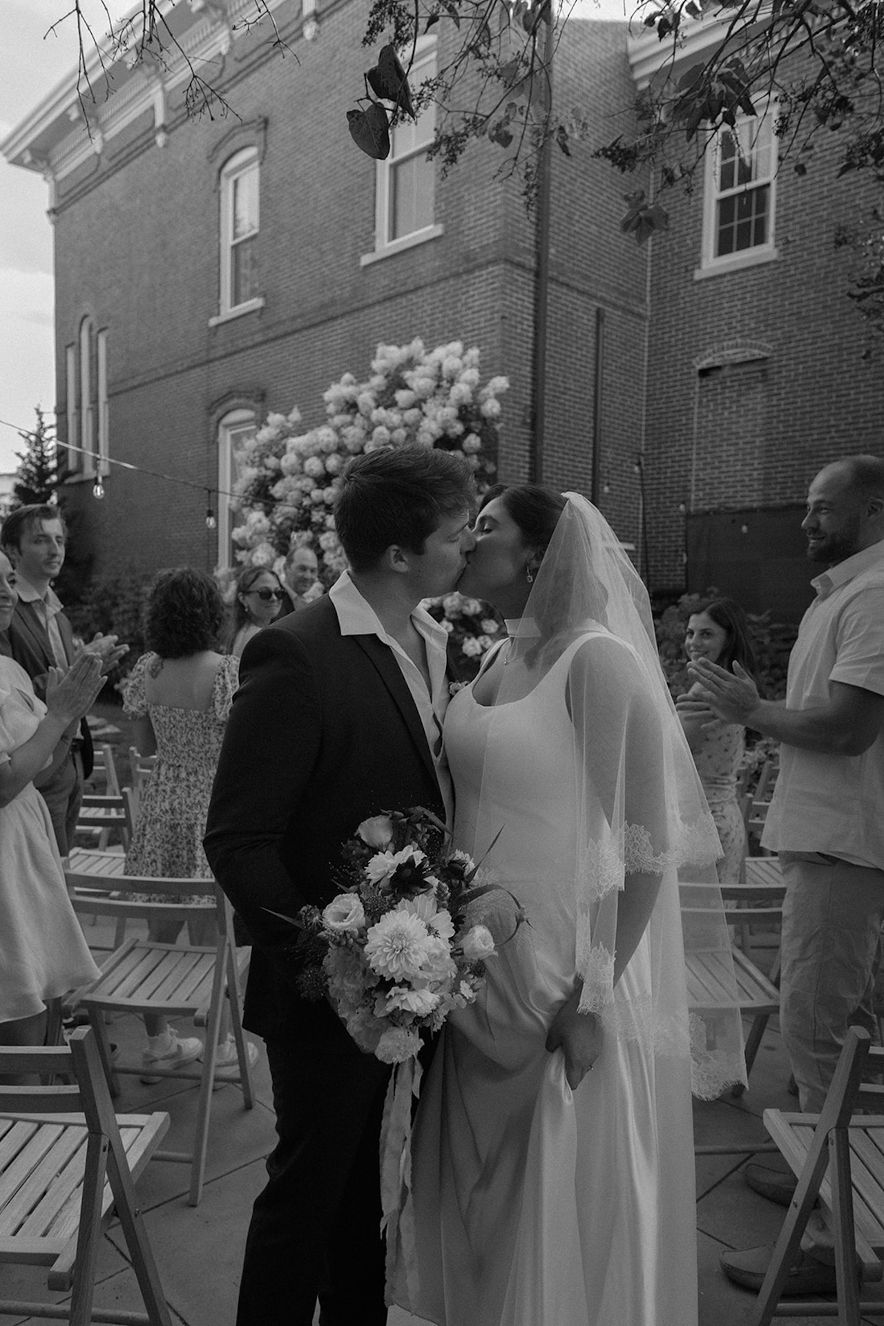 Black-and-white photo of a bride and groom kissing after their ceremony, surrounded by clapping guests and lush florals, highlighting Destination Wedding Location Ideas.