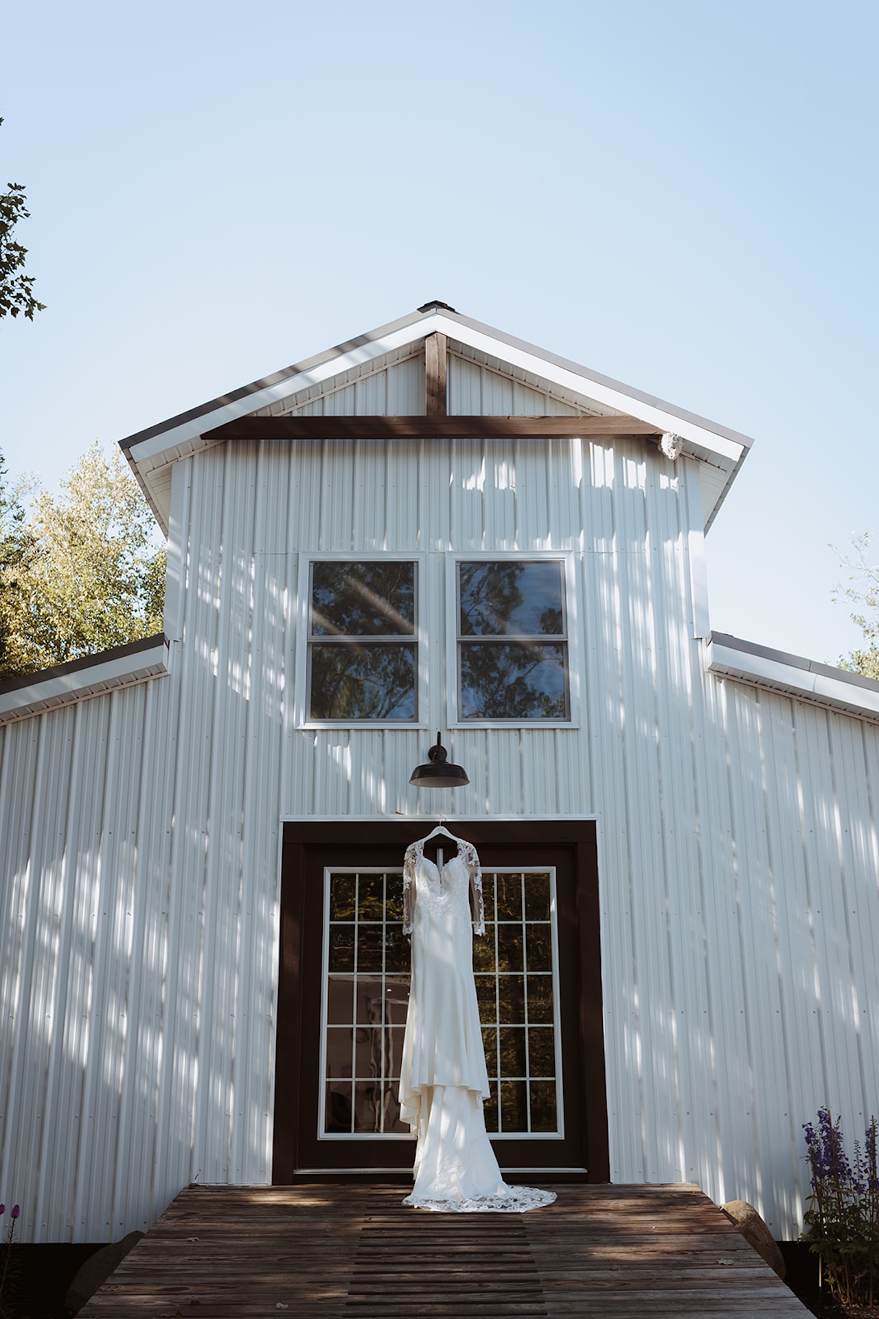 A wedding dress with lace sleeves hanging on a barn door, sunlight casting shadows on the white siding, rustic and elegant Destination Wedding Location Ideas.