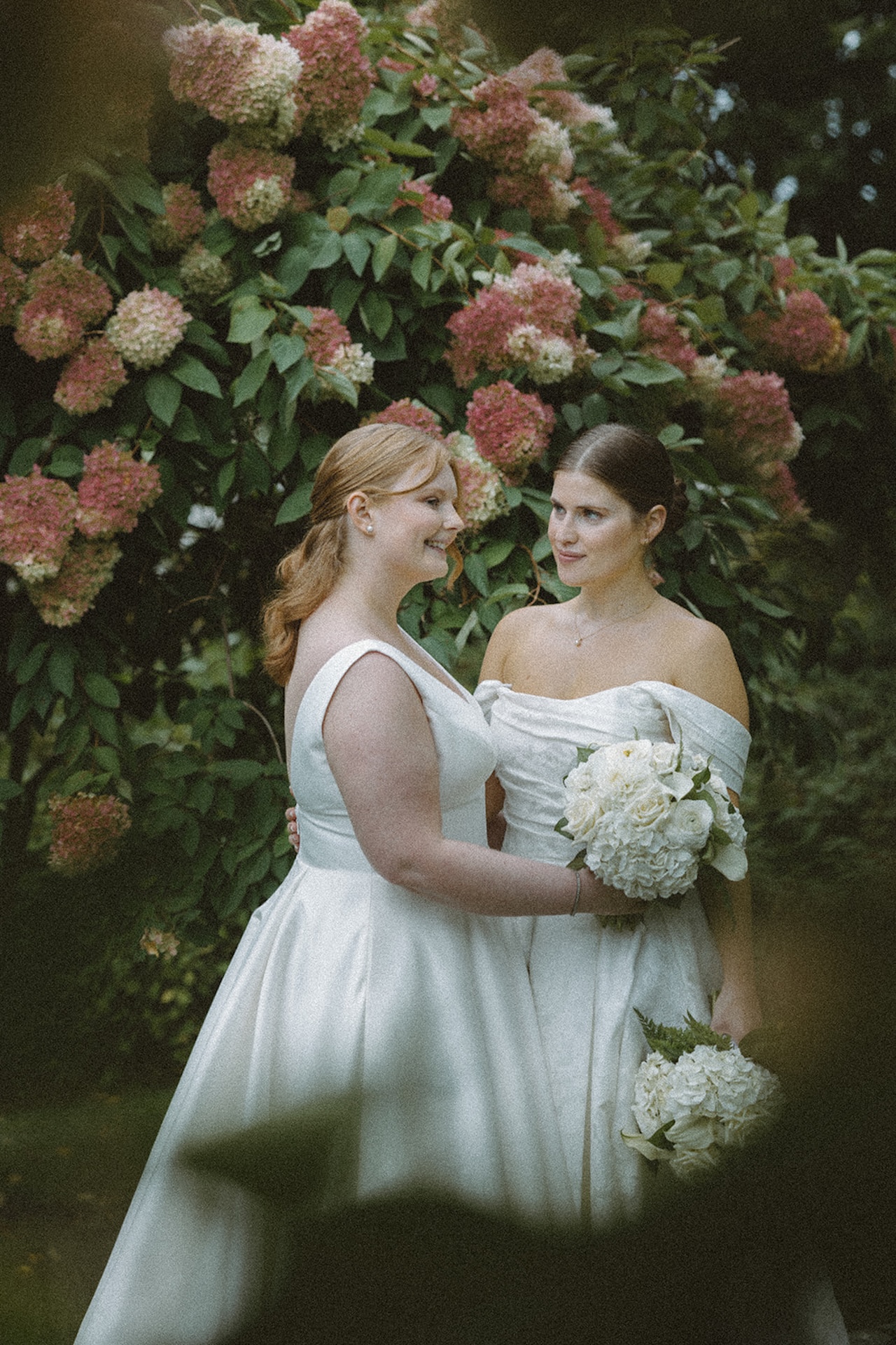 Two brides standing together in front of blooming hydrangeas, holding bouquets and sharing a quiet, joyful moment.
