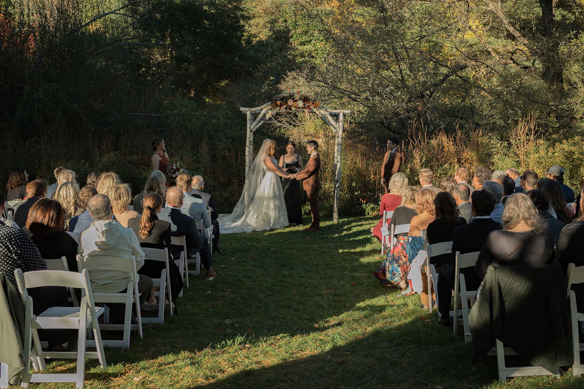 Outdoor wedding ceremony with a rustic birch arch, couple exchanging vows surrounded by family and friends in a natural setting.