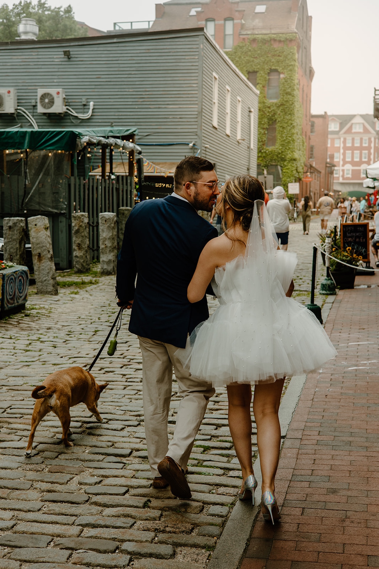 Couple walking their dog down cobblestone streets after their ceremony, a candid and playful wedding day stroll.
