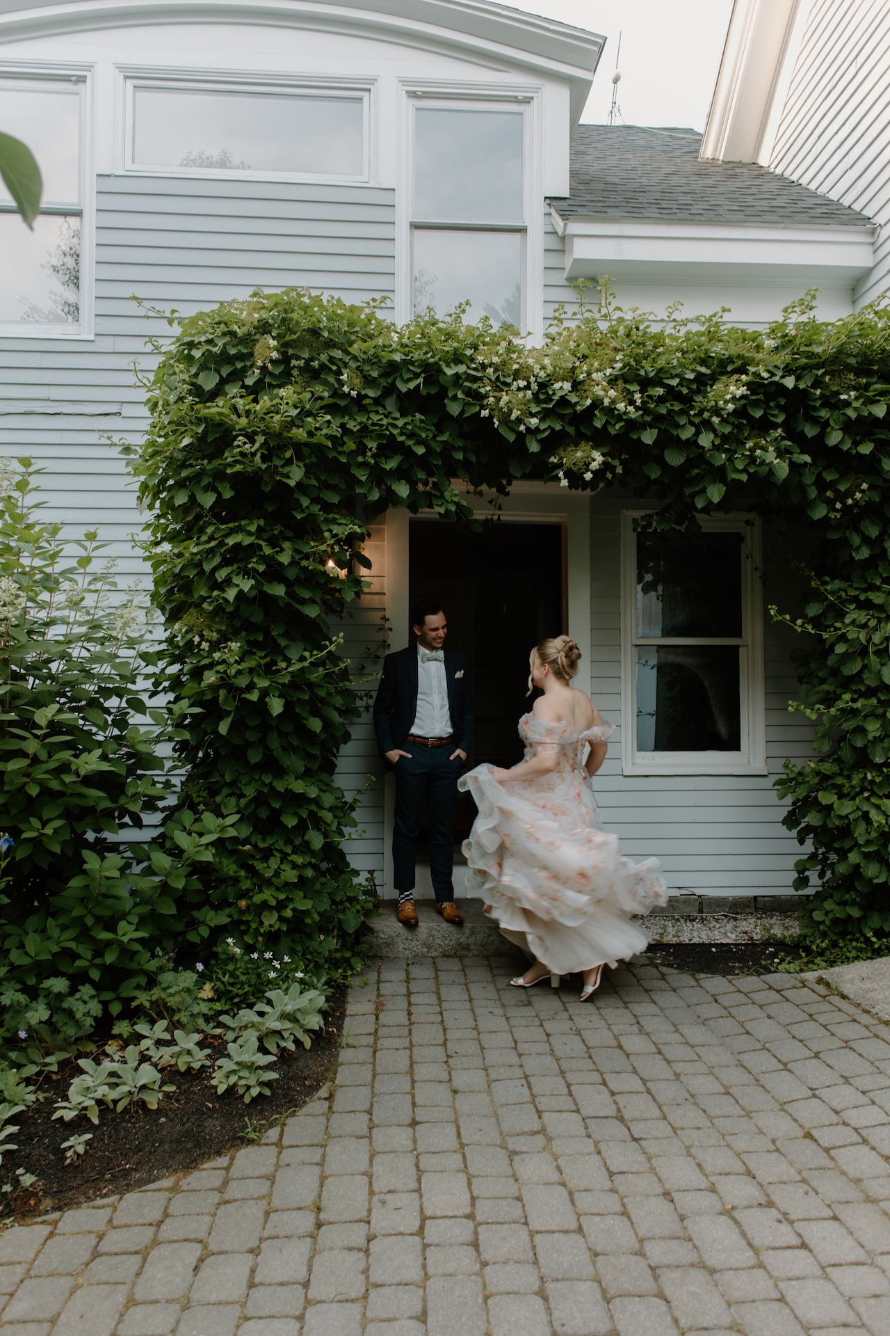 A candid moment of the groom watching his bride twirl in her layered floral gown, framed by ivy-covered walls of a farmhouse.