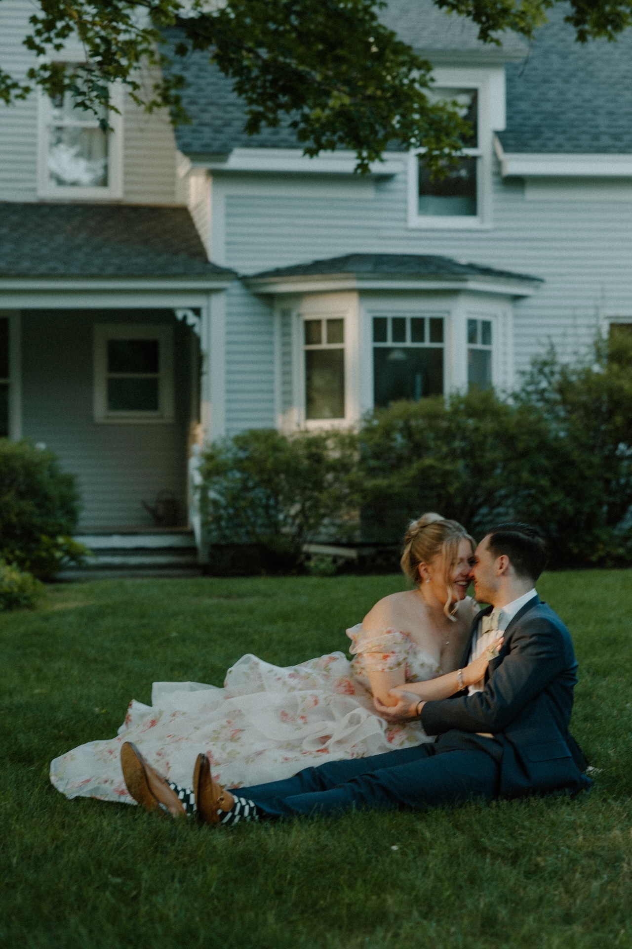 The bride and groom sitting close on the lawn, laughing together as the evening light glows at a Portland Maine wedding venue.