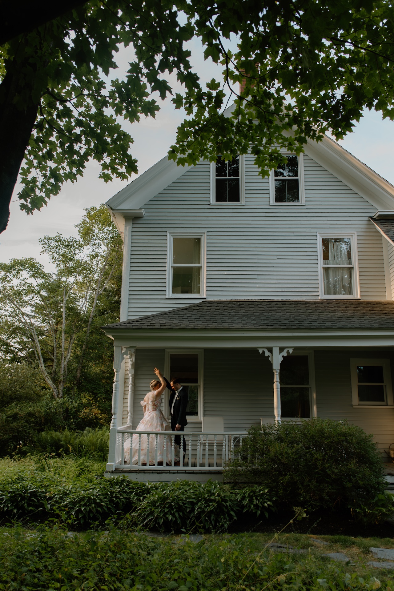The couple dancing together on the porch of a classic New England-style home, framed by leafy branches.