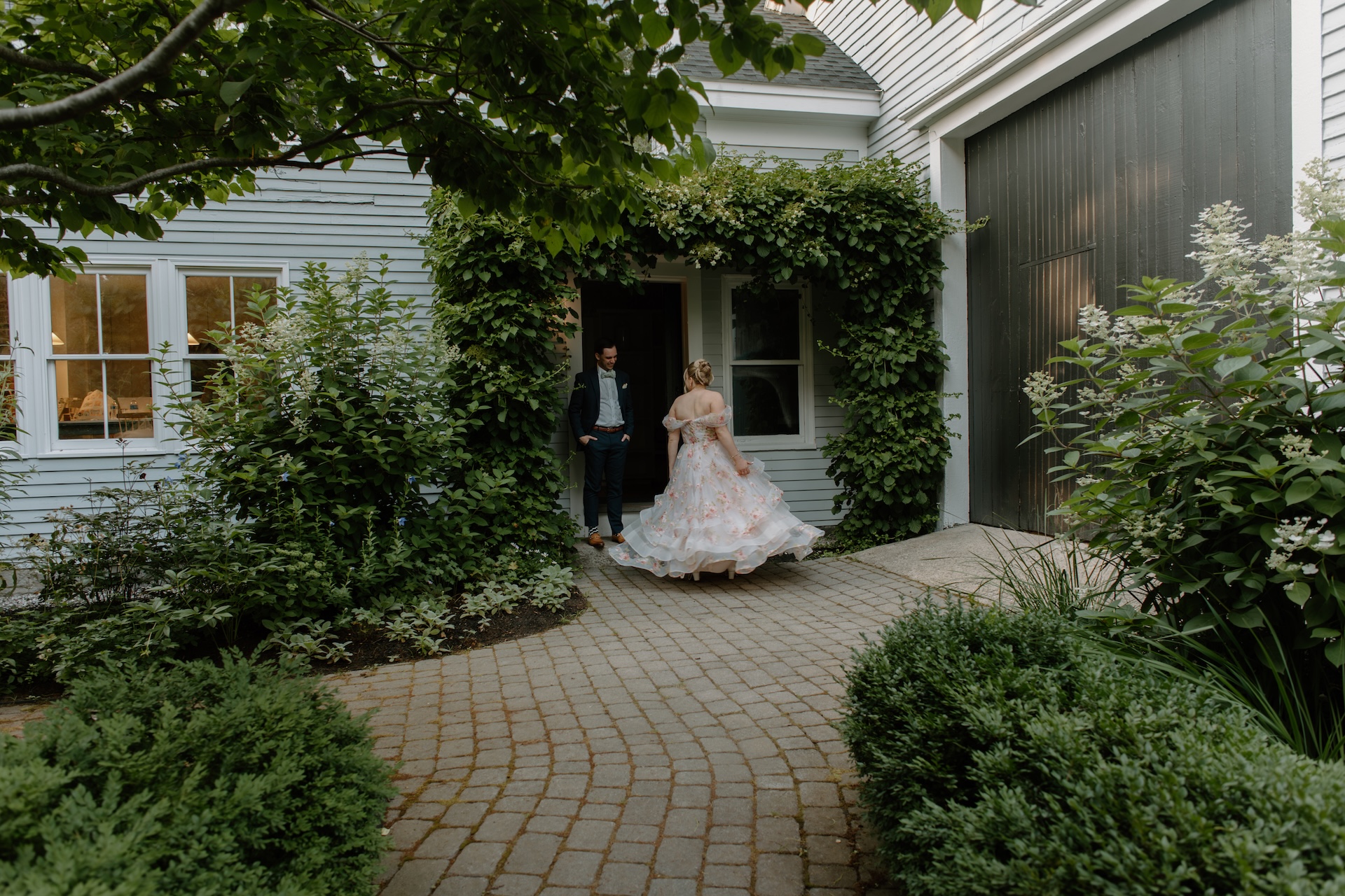 The bride twirling her floral gown in front of the farmhouse while the groom watches, framed by lush greenery.