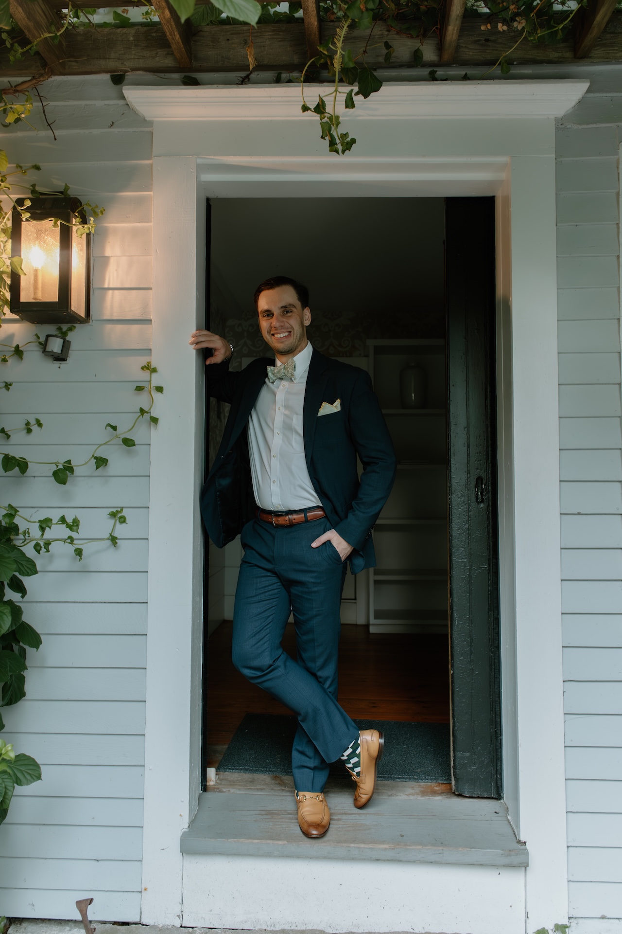 The groom leaning casually in a farmhouse doorway, dressed in a navy suit with a bow tie and loafers.