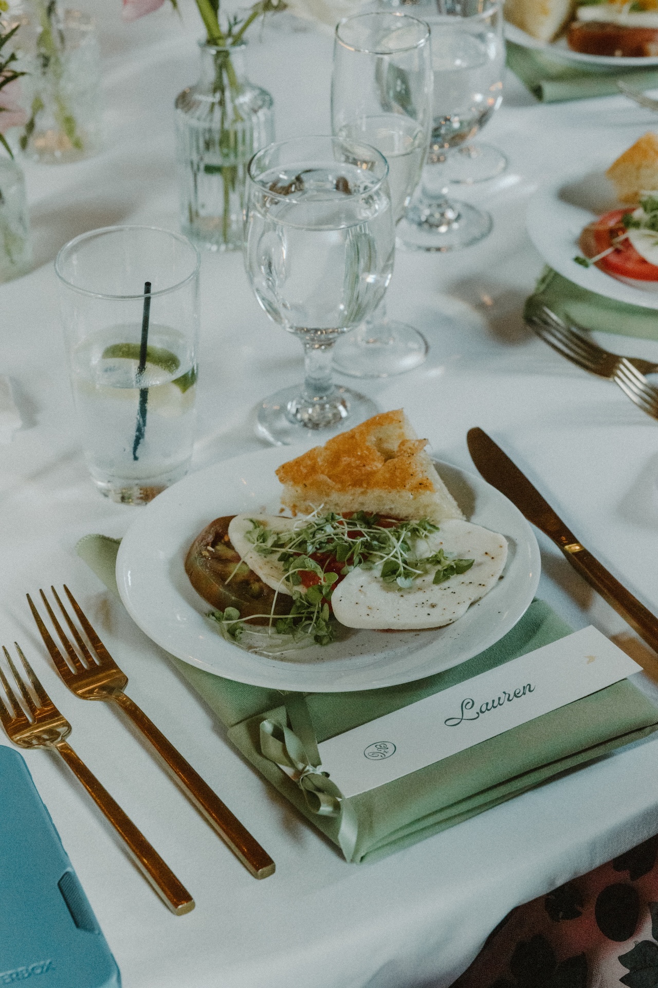 A close-up of a plated dinner salad with mozzarella, tomato, and focaccia bread, placed neatly beside gold flatware.