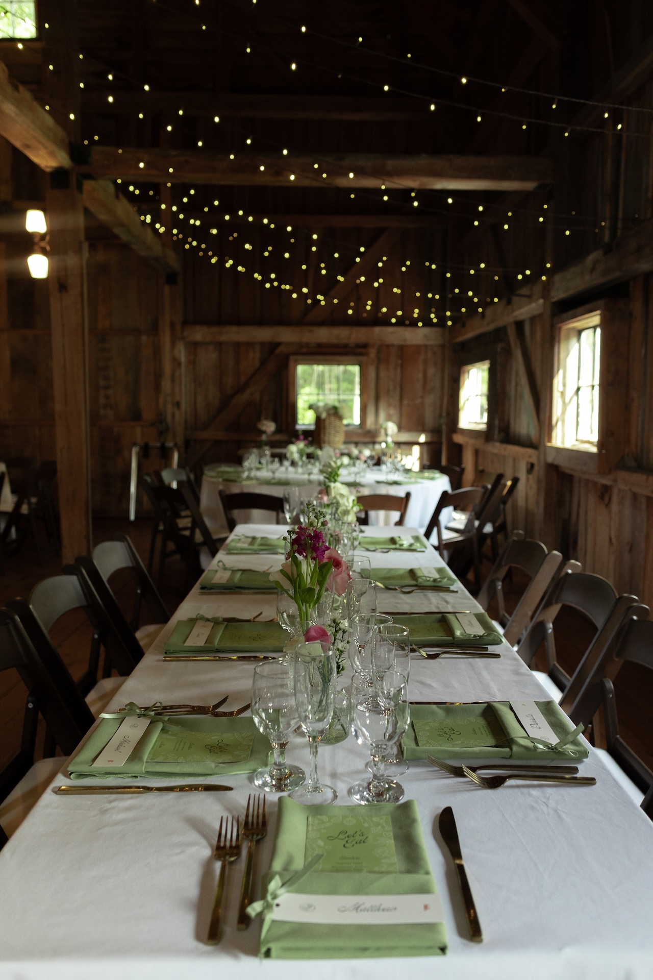 Reception tables decorated with green napkins, glassware, and twinkling lights strung across the rustic barn beams.