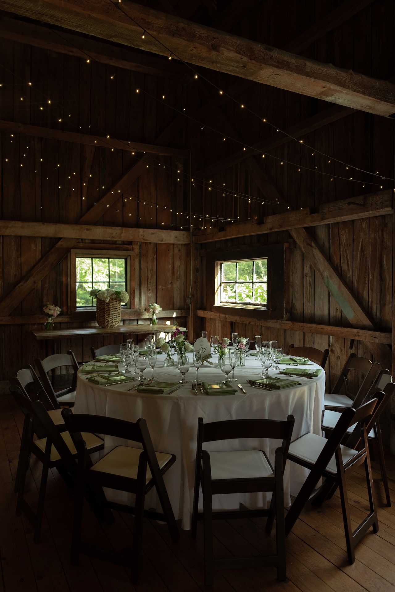Reception setup inside the barn with round tables decorated in white linens, green napkins, glassware, and twinkle lights at a Portland Maine wedding venue.