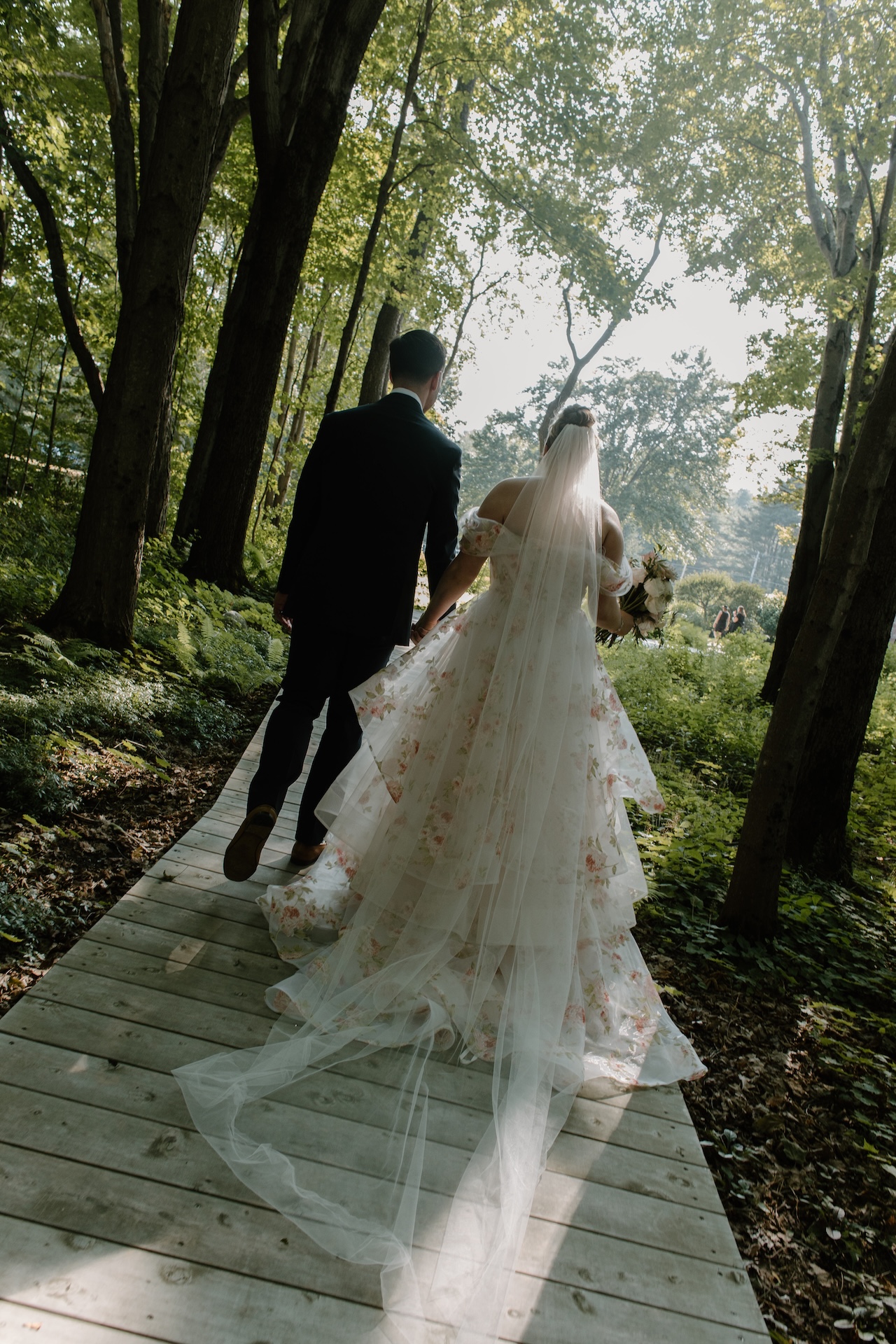 The bride and groom walking hand in hand down a wooden path surrounded by tall trees at a Portland Maine wedding venue.