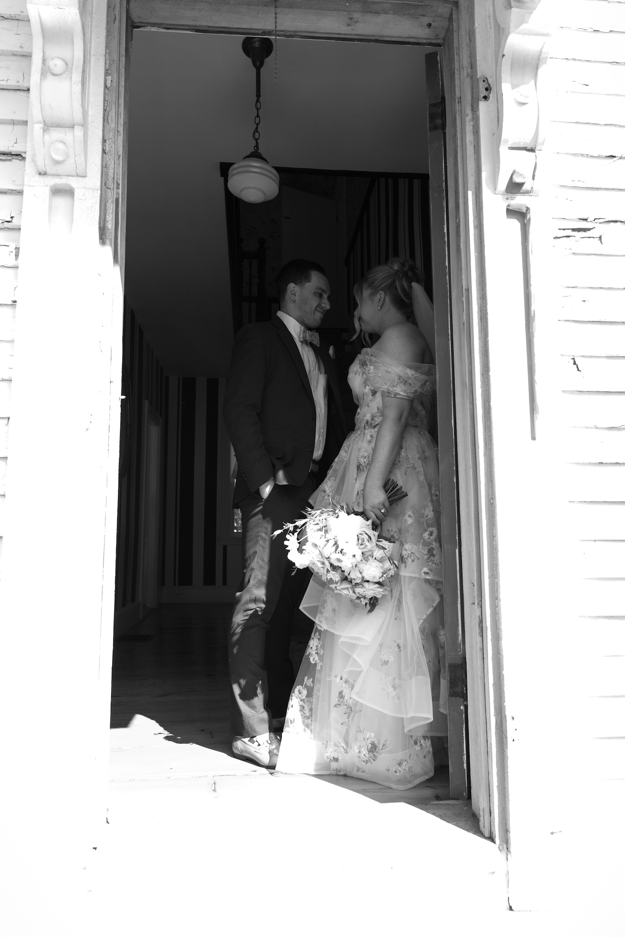 A black-and-white photo of the bride and groom standing in the farmhouse doorway, gazing at each other at a Portland Maine wedding venue.