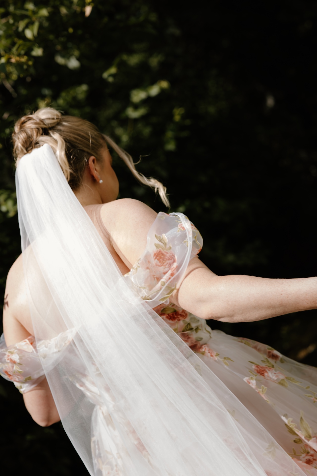 A close-up from behind the bride, showing her flowing veil and romantic floral dress as sunlight filters through the trees.