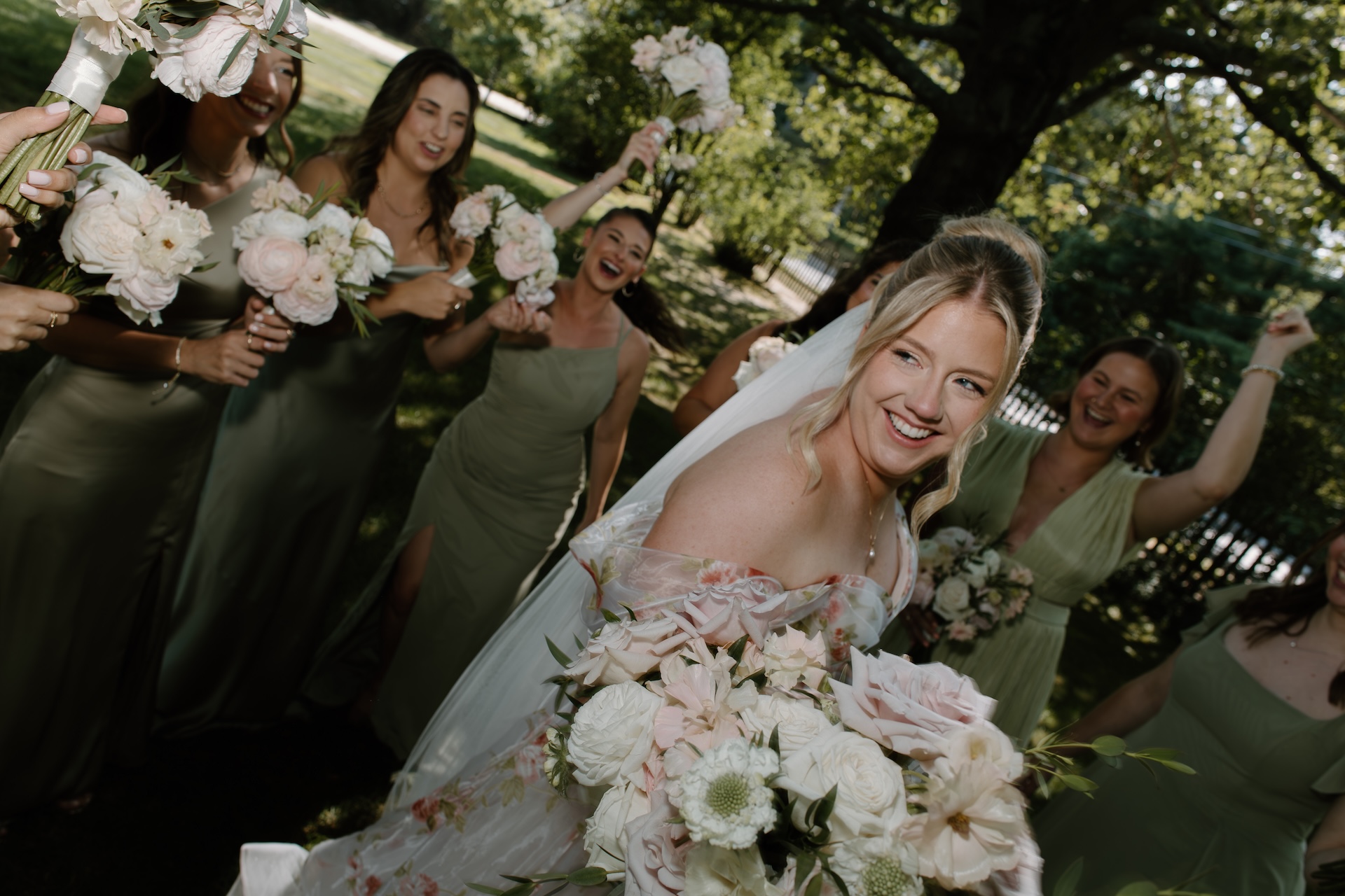 The bride smiling brightly while surrounded by bridesmaids in sage dresses, everyone cheering with bouquets in hand.