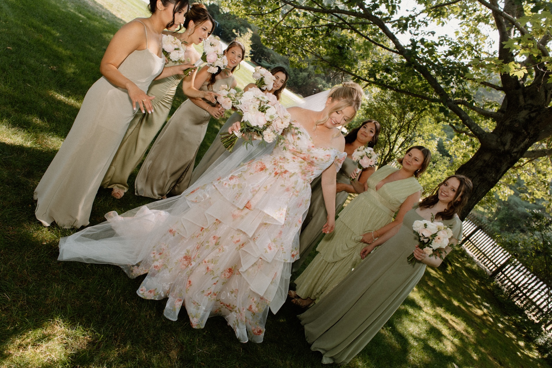 The bride smiling while walking hand-in-hand with her bridesmaids in sage green dresses, all holding soft blush bouquets at a Portland Maine wedding venue.