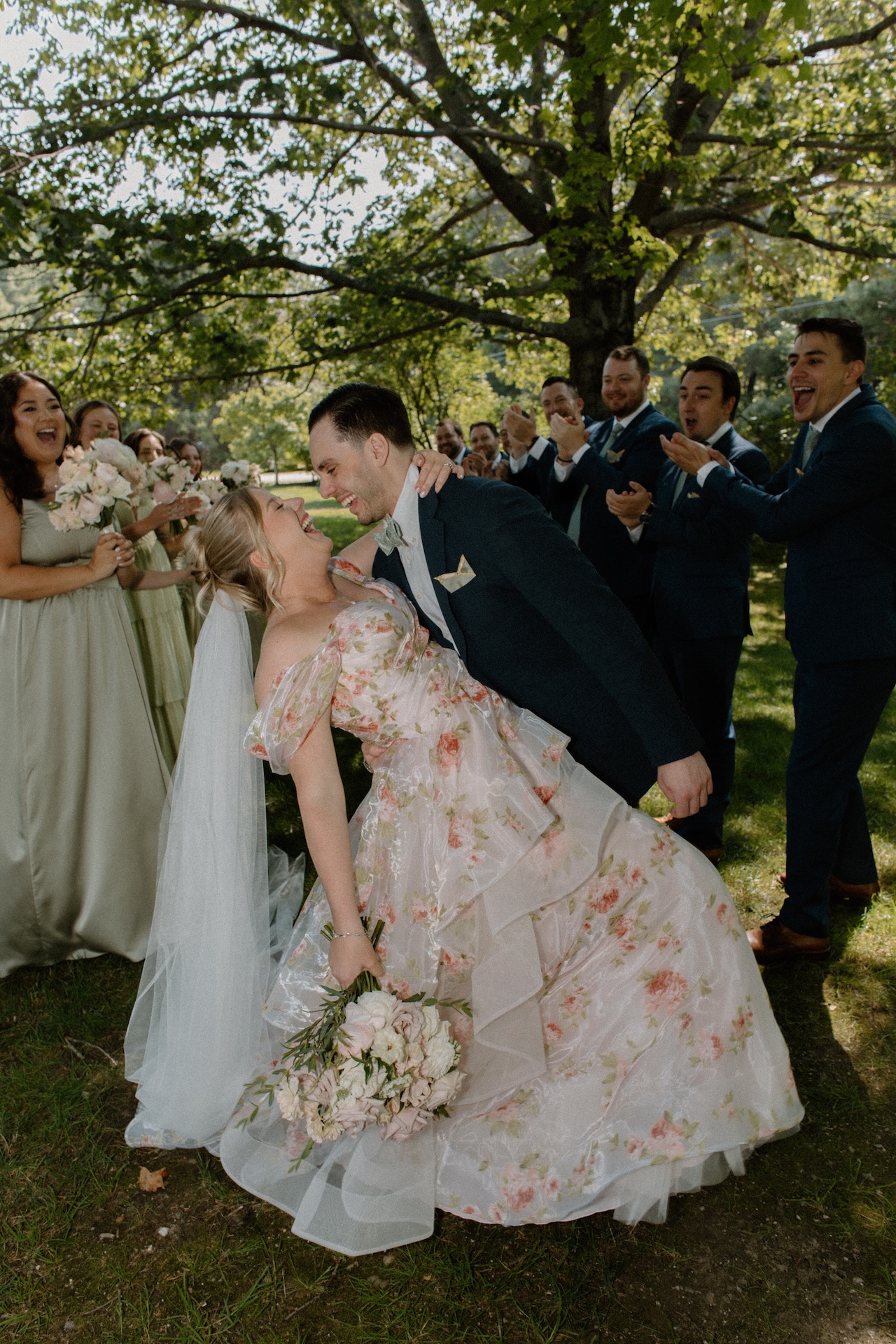 The bride in her floral gown and the groom in a navy suit sharing a playful dip kiss, surrounded by their cheering wedding party at a Portland Maine wedding venue.