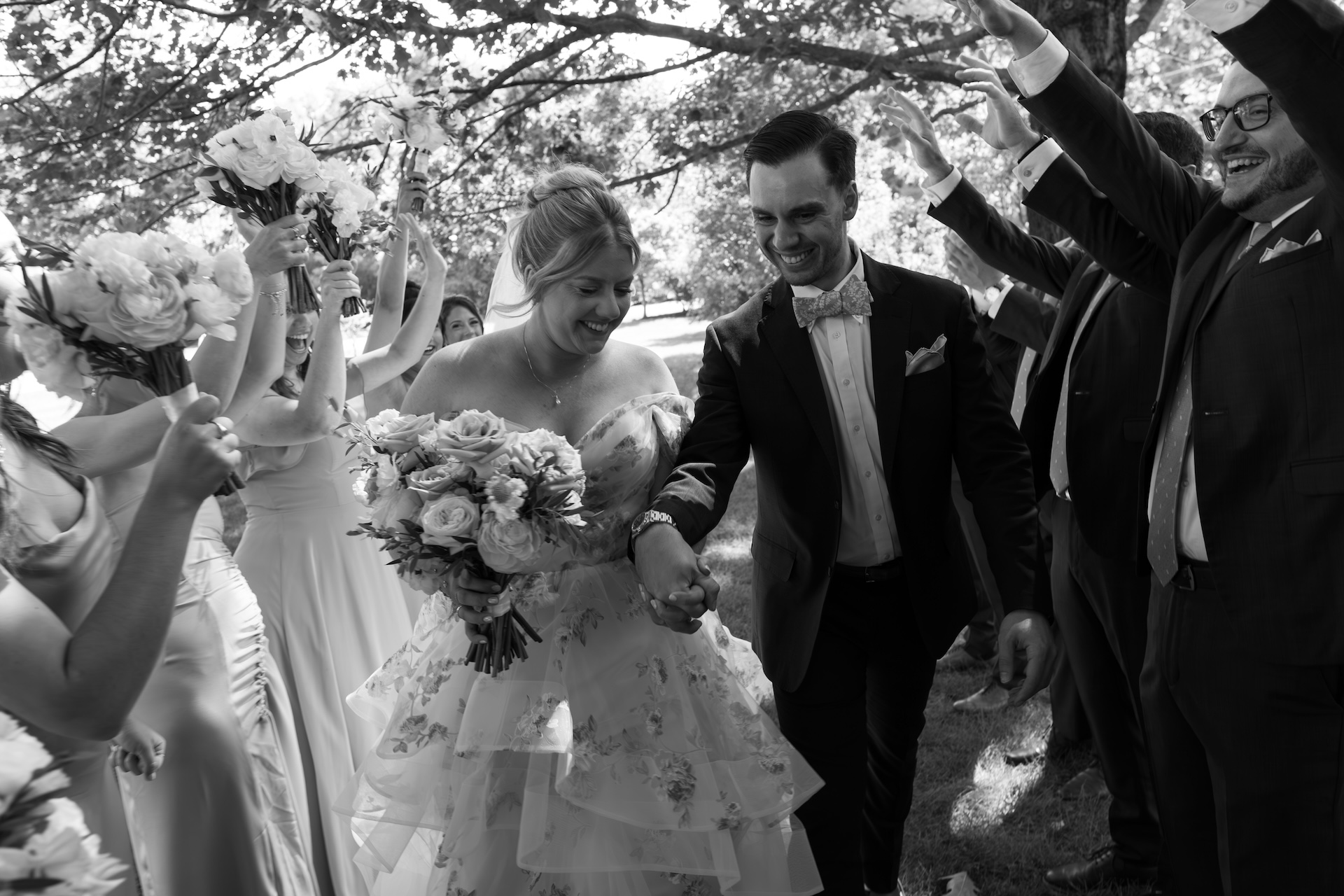 A black-and-white photo of the bride and groom walking through a tunnel of cheering friends holding bouquets.