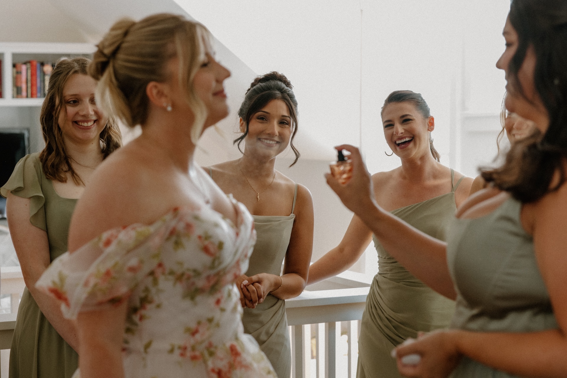 Bridesmaids in sage gowns spraying perfume on the bride as everyone laughs together during morning preparations.