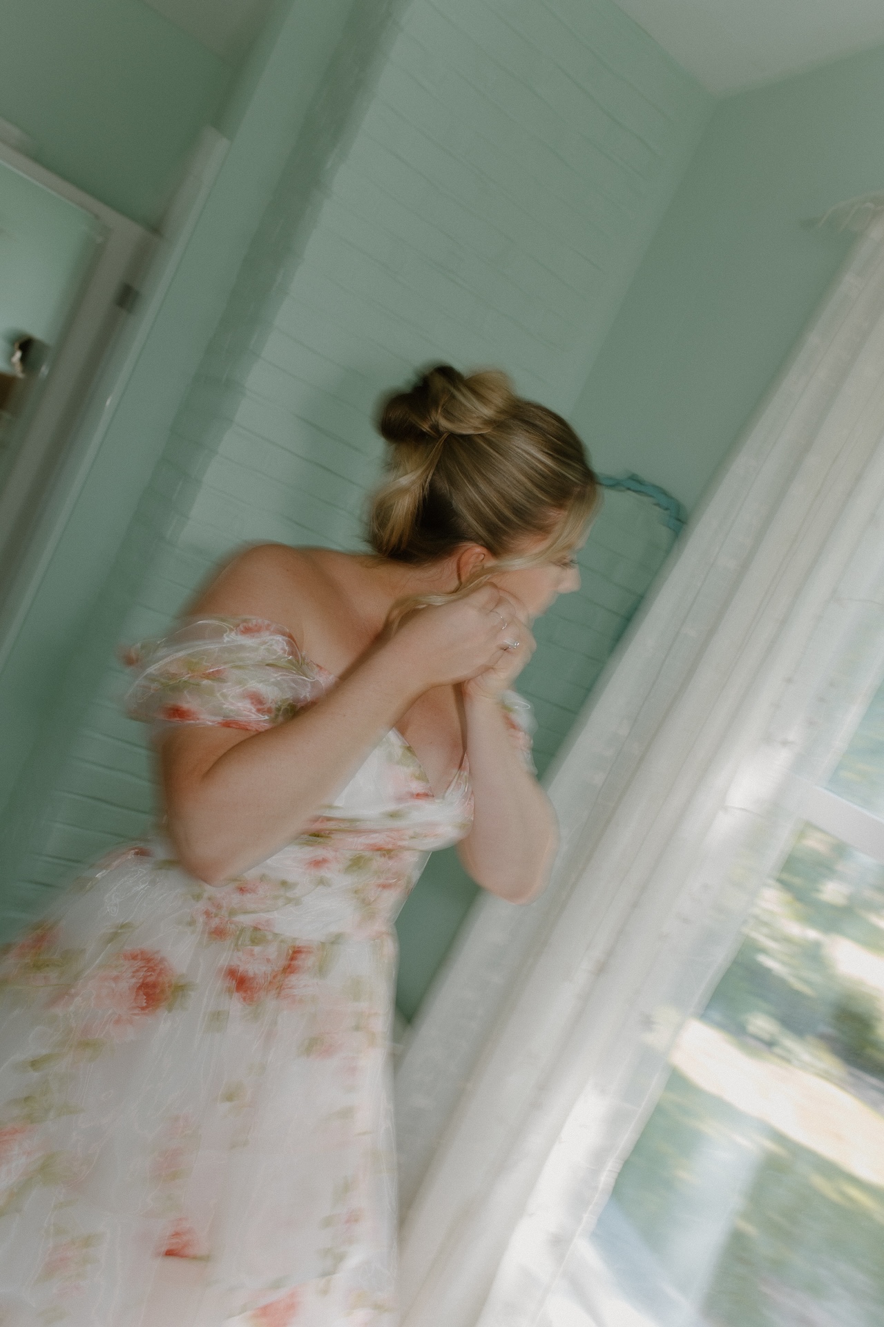 The bride adjusting her earring by a sunlit window, her floral gown glowing softly in the natural light at a Portland Maine wedding venue.
