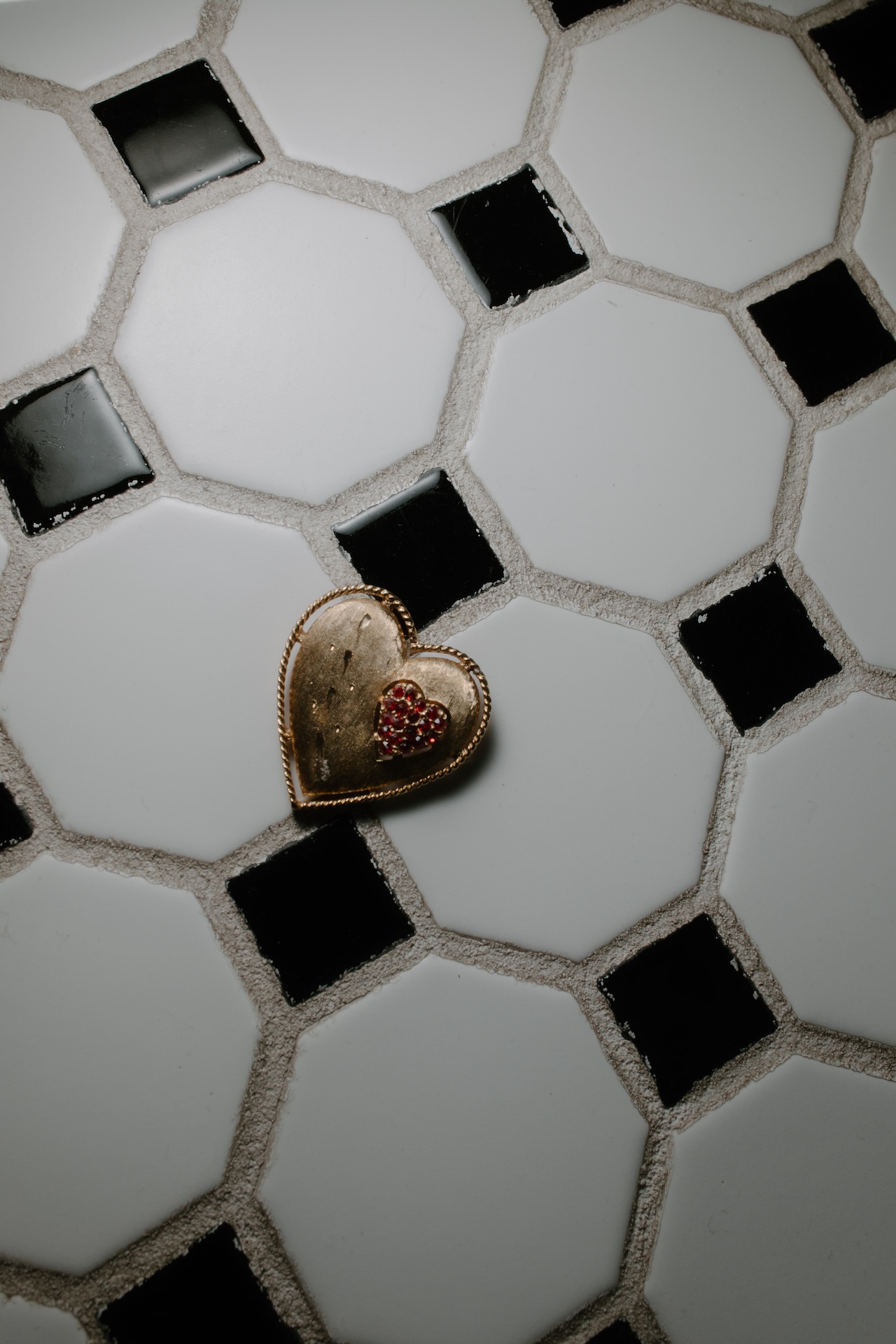 A small gold heart-shaped locket with a red jeweled heart accent resting on black-and-white tile flooring.