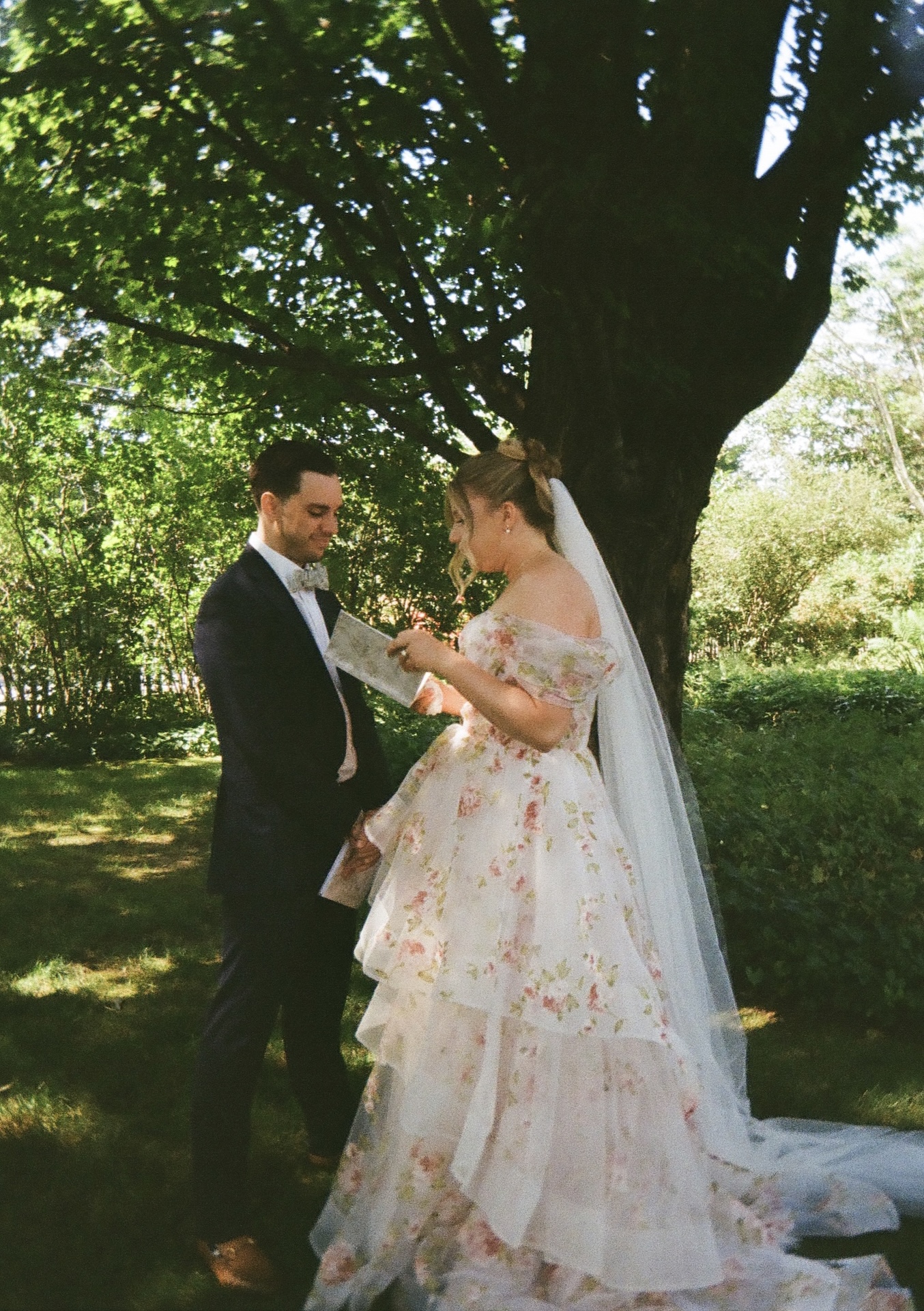 The bride and groom exchanging handwritten vows beneath the shade of tall trees at a Portland Maine wedding venue.