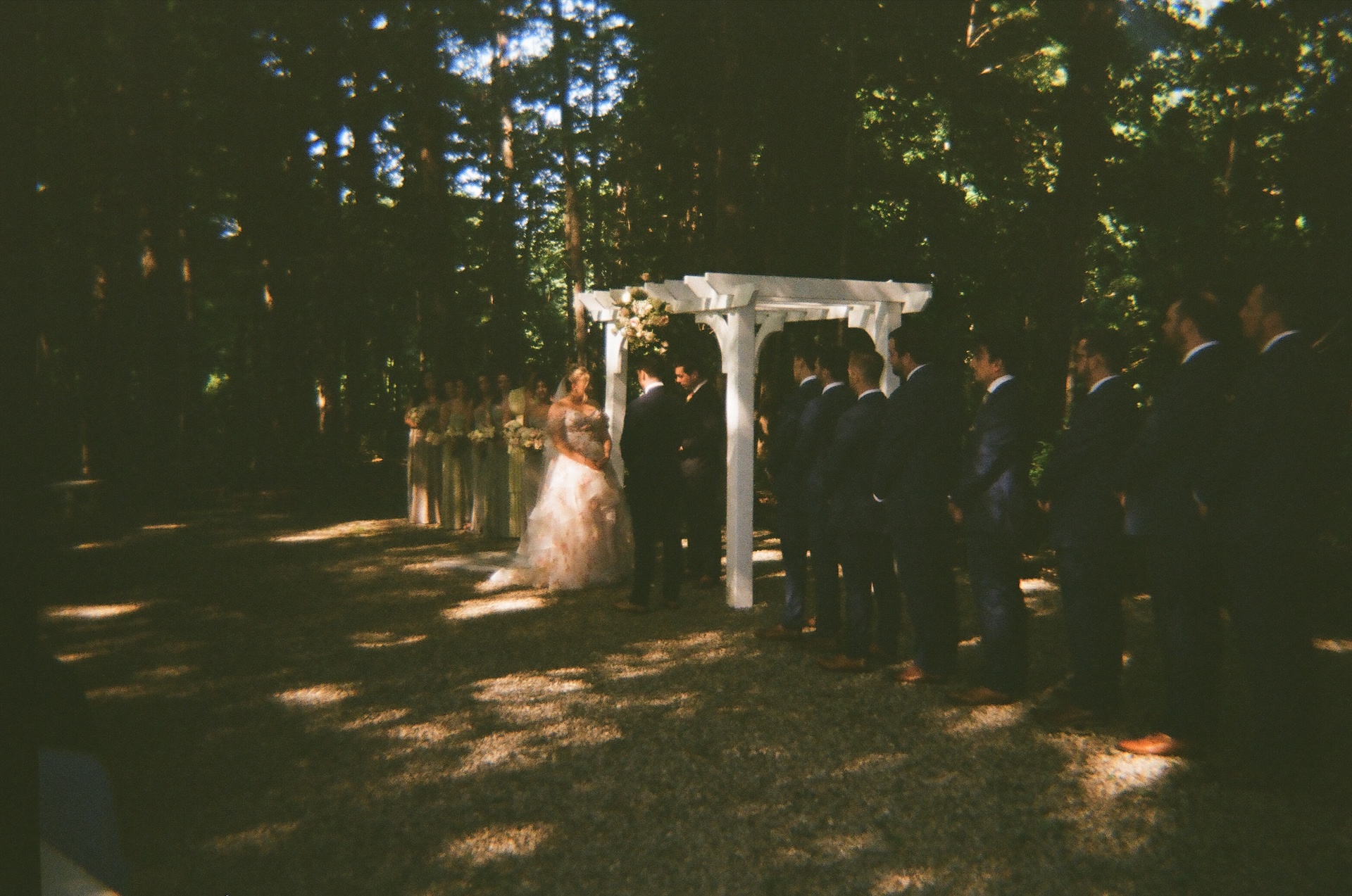 The couple standing under a white wooden arbor as they exchange vows, surrounded by their wedding party at a Portland Maine wedding venue.