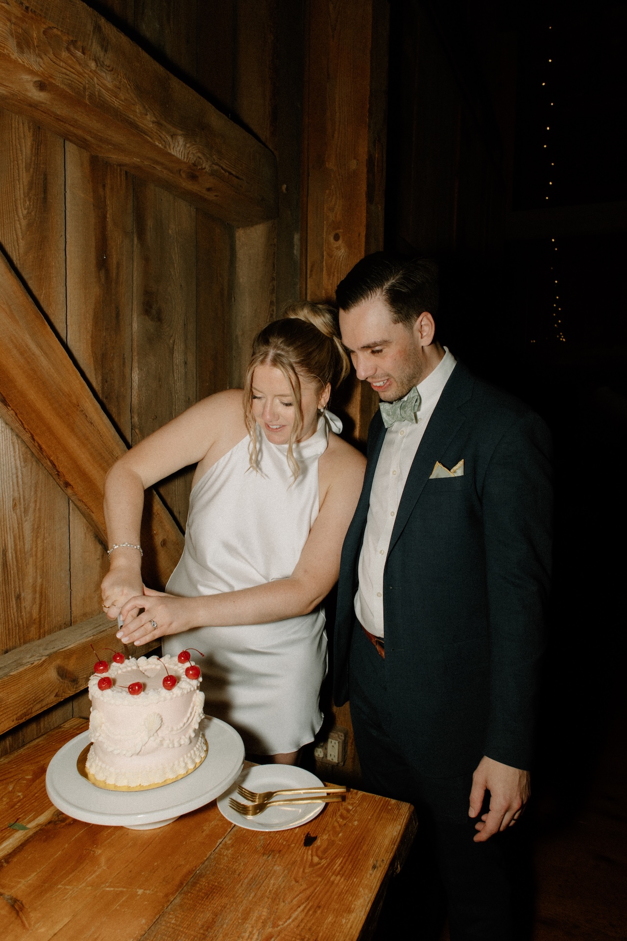 The bride and groom cutting their cherry-topped cake together inside the rustic barn, smiling as they celebrate.