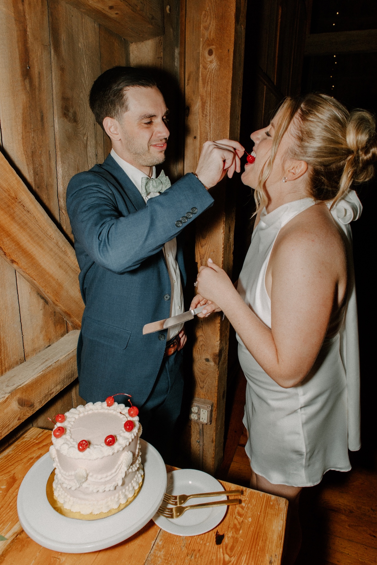 The groom feeding the bride a cherry from their vintage-inspired wedding cake inside the barn at a Portland Maine wedding venue.