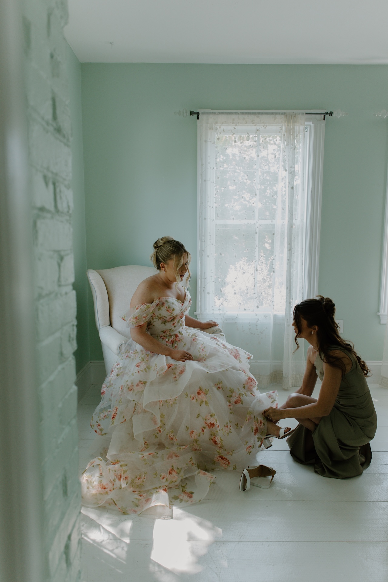 A bridesmaid helping the bride with her shoes in a sunlit getting-ready room painted soft green at a Portland Maine wedding venue.