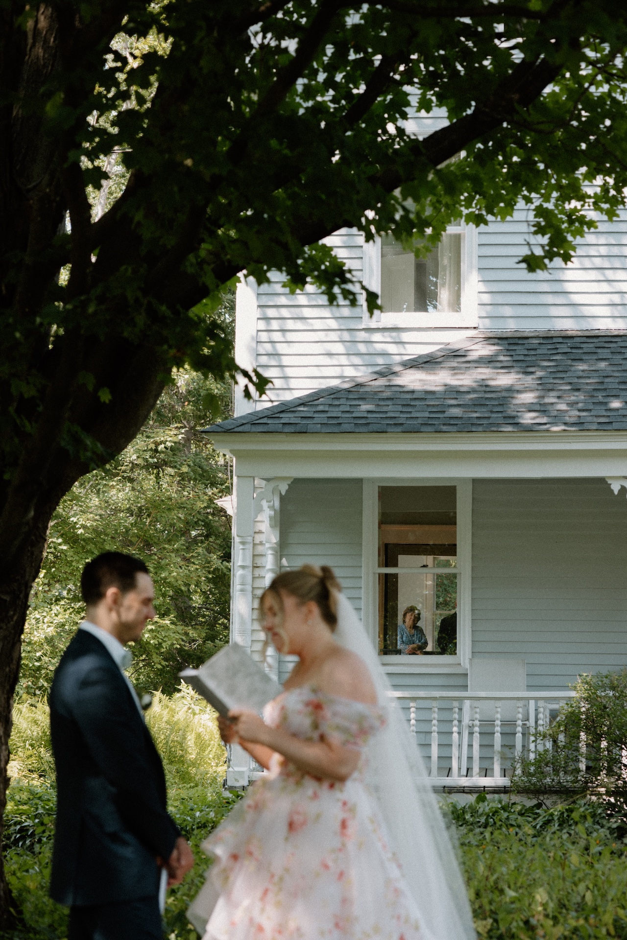 The bride reading vows to the groom in the garden, while a family member watches tenderly through a farmhouse window at a Portland Maine wedding venue.
