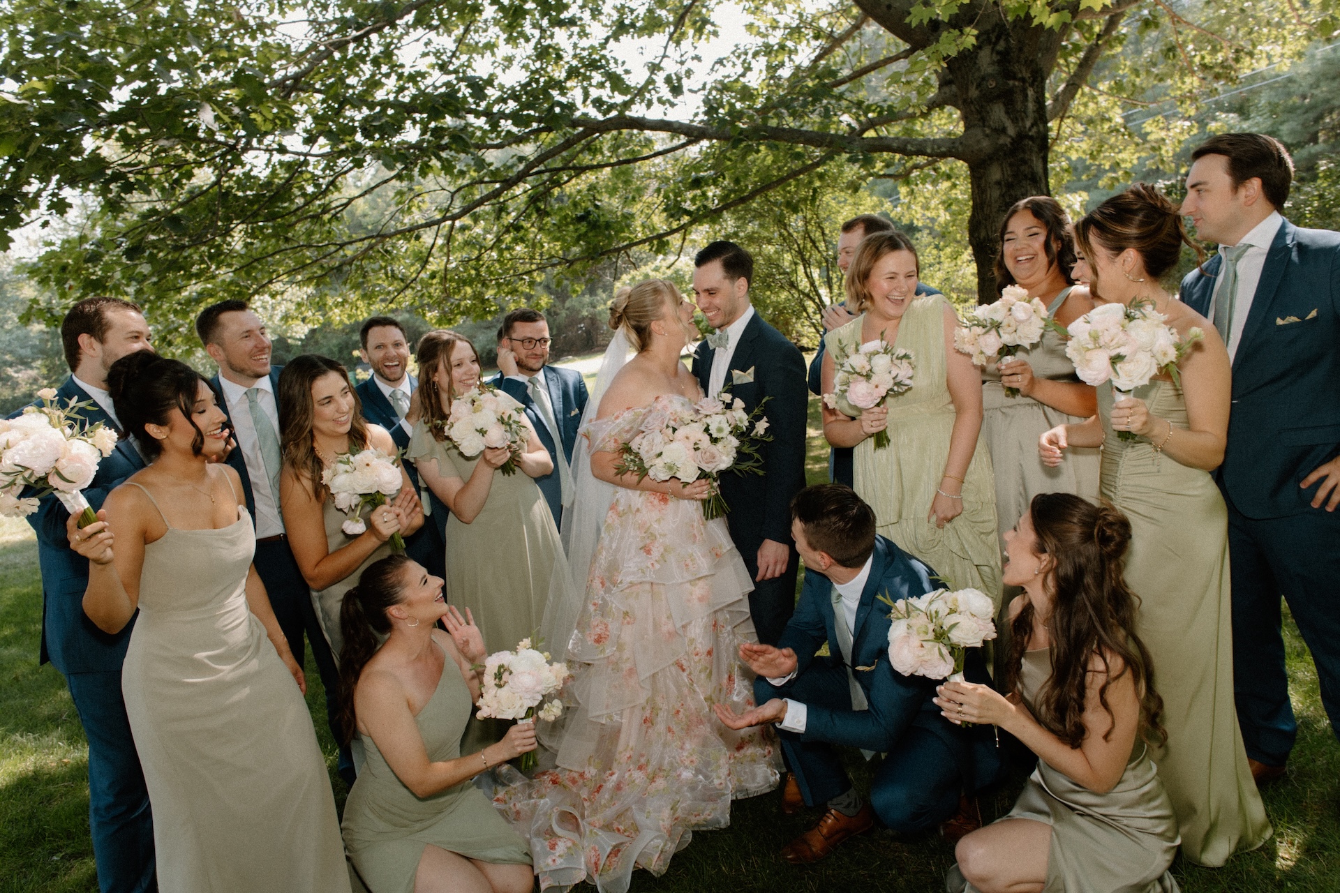 A joyful group shot of the bride and groom laughing with bridesmaids and groomsmen, all gathered under a large tree.