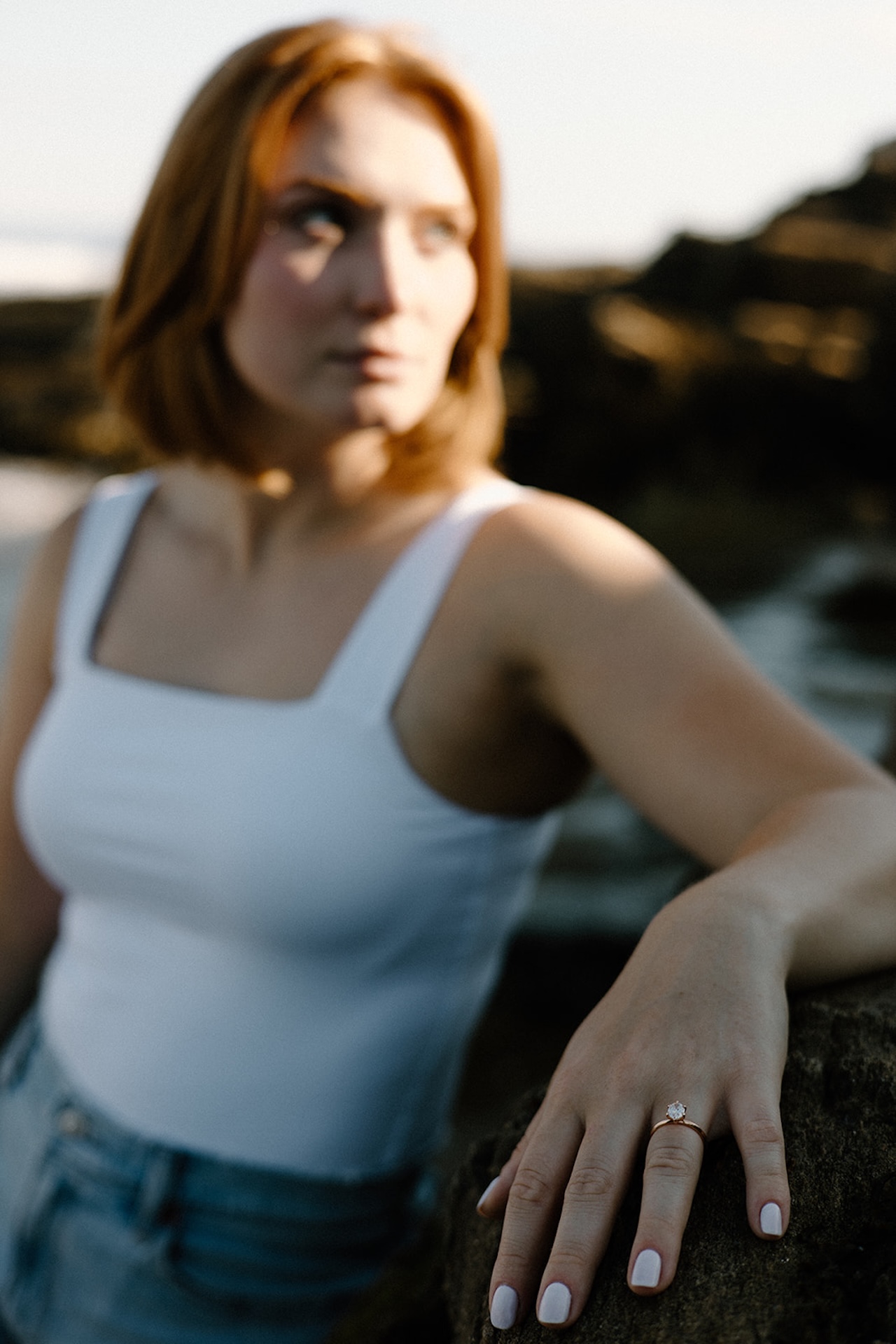 Close-up of a woman’s hand resting on a rock, showing her engagement ring with ocean light behind her.