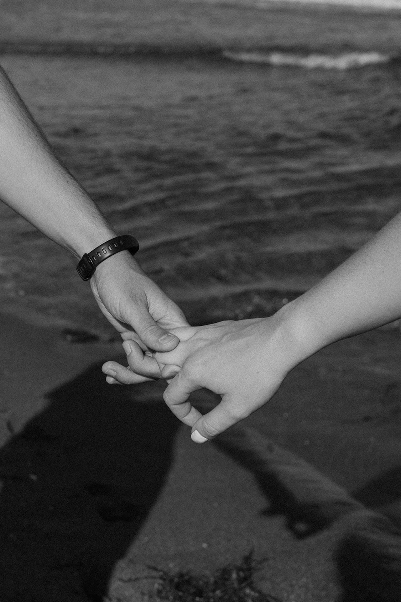 Black and white photo of hands intertwining on the beach, waves softly blurring in the background.