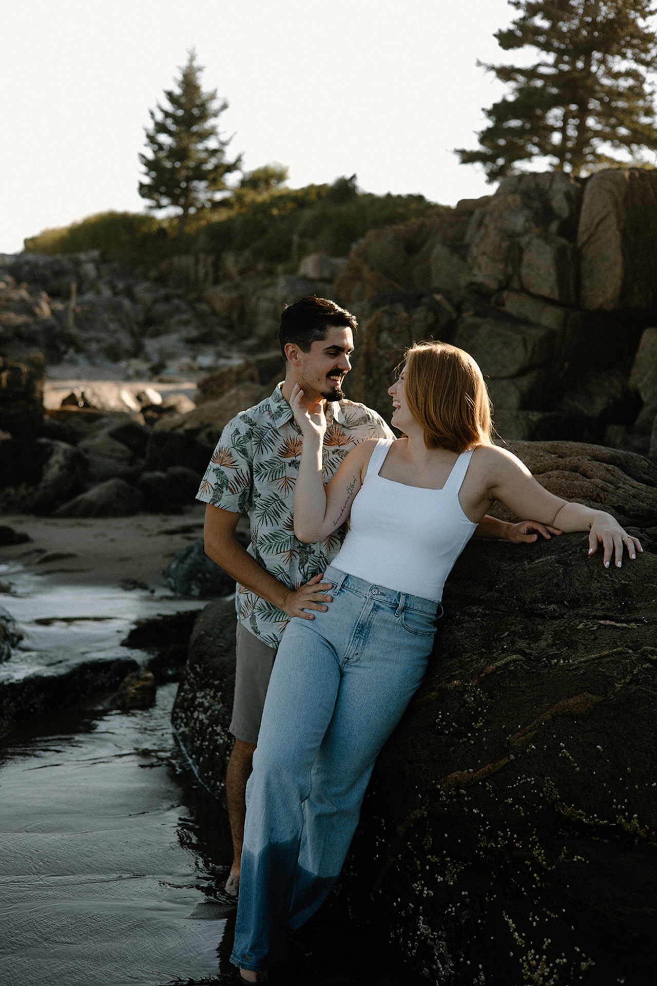 Couple laughing together on rocky shoreline, dressed casually for a coastal evening shoot.
