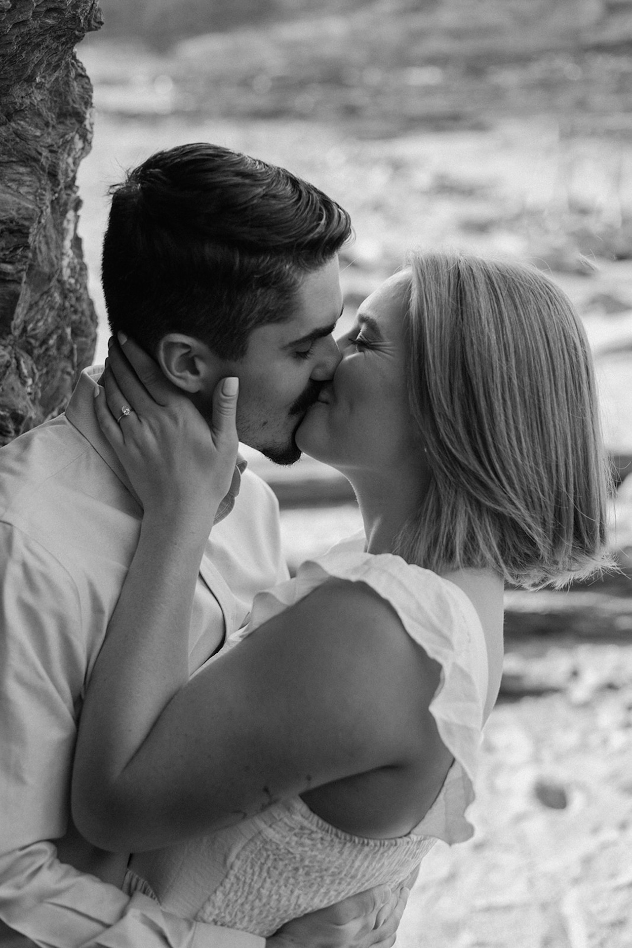 Couple sharing a kiss beside a rocky shoreline, the sunlight softly illuminating their faces.