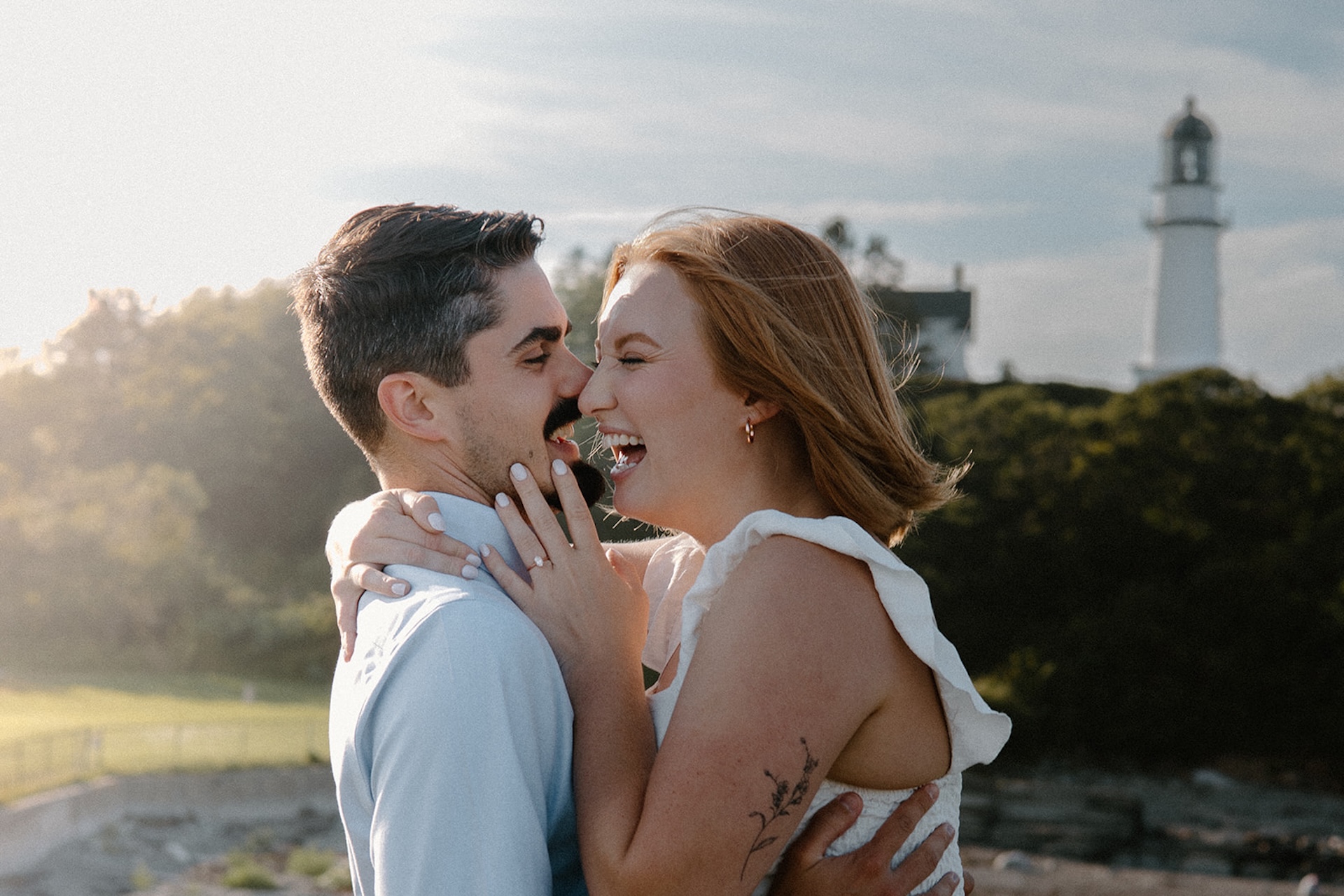 Couple laughing together in front of a lighthouse during golden hour, a joyful moment captured at one of Maine’s most romantic Engagement Photo Locations.