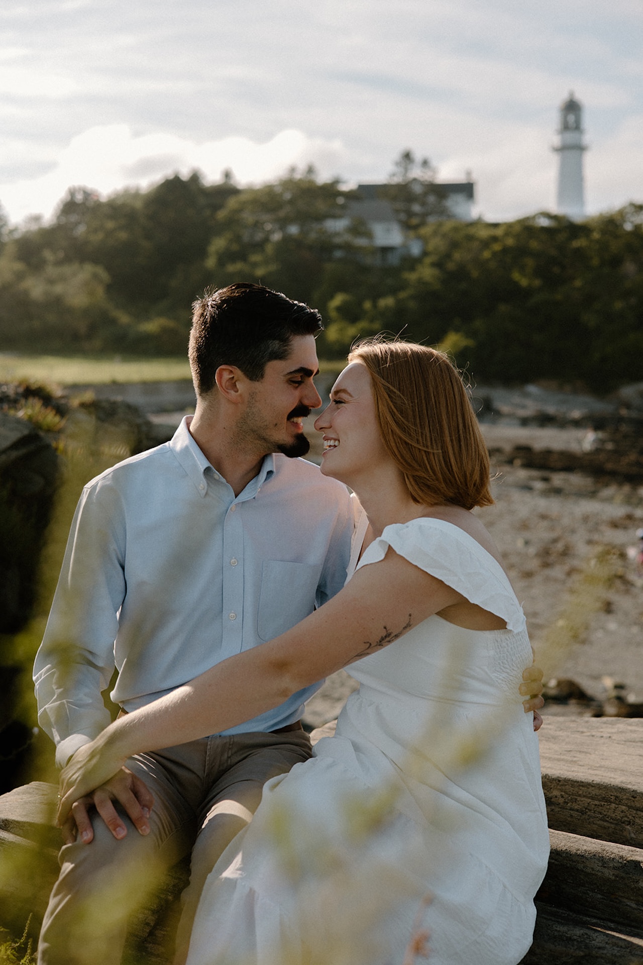 Couple sitting on driftwood by the water, smiling as they lean in close under the warm evening light.